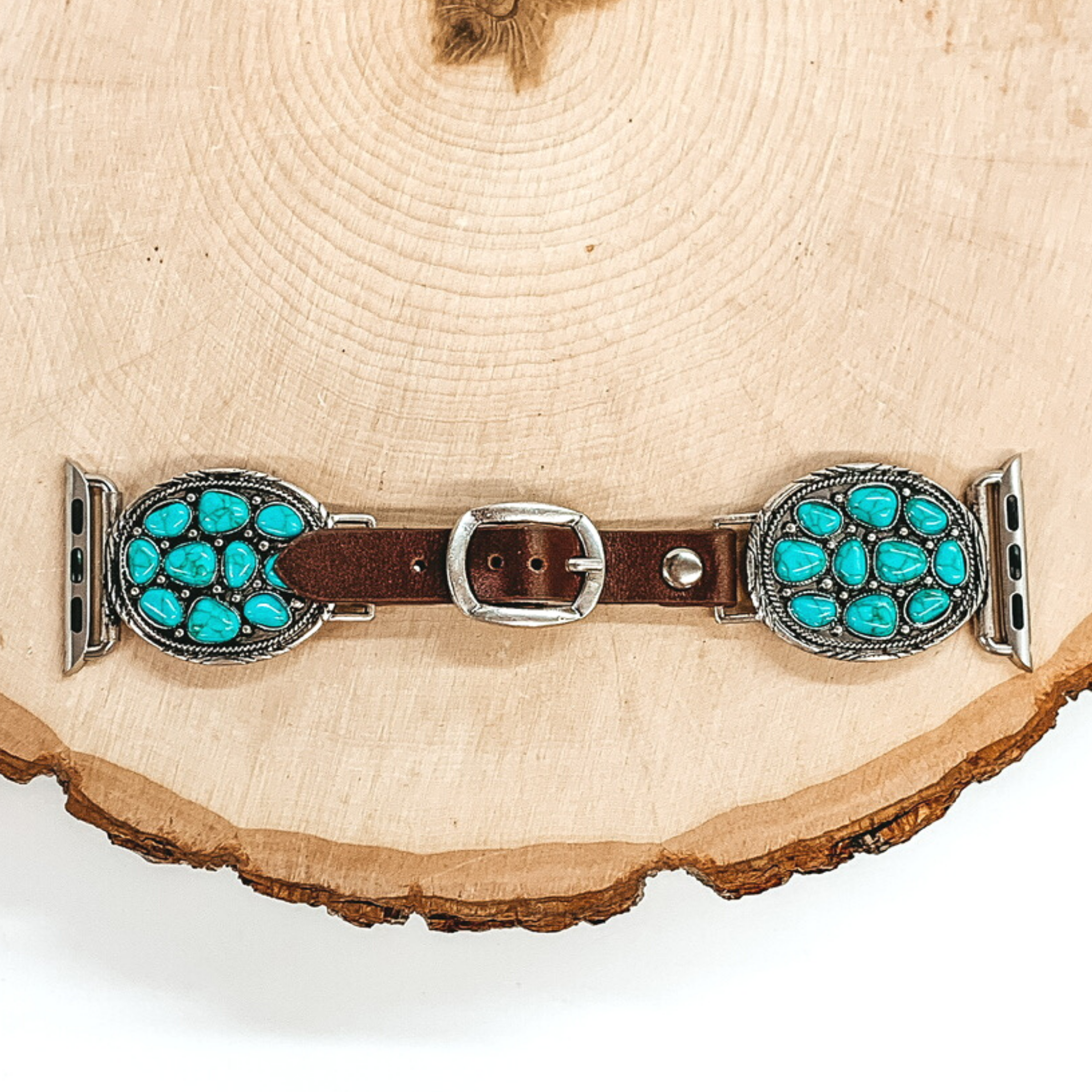 Dark Brown watch band with silver, oval pendant ends with Apple watch band acessories. The oval pendant has irregular shaped turquoise stones. This watch band is pictured on a piece of wood on a white background. 