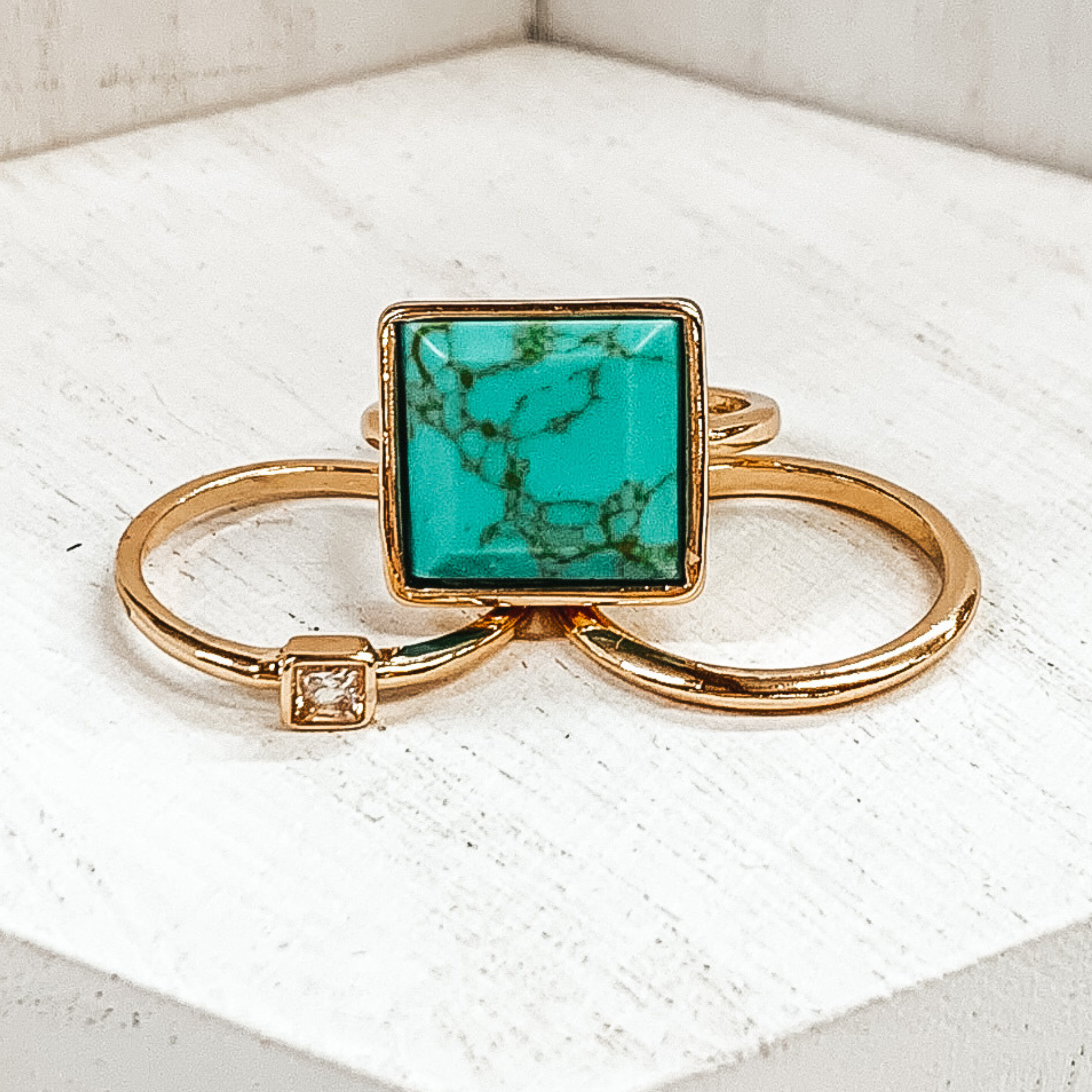 Set of three gold rings. One ring is a plain gold band. Another has a small square pendant with a center crystal. The third ring has a big, turquoise pendant. These rings are pictured on a white background.  