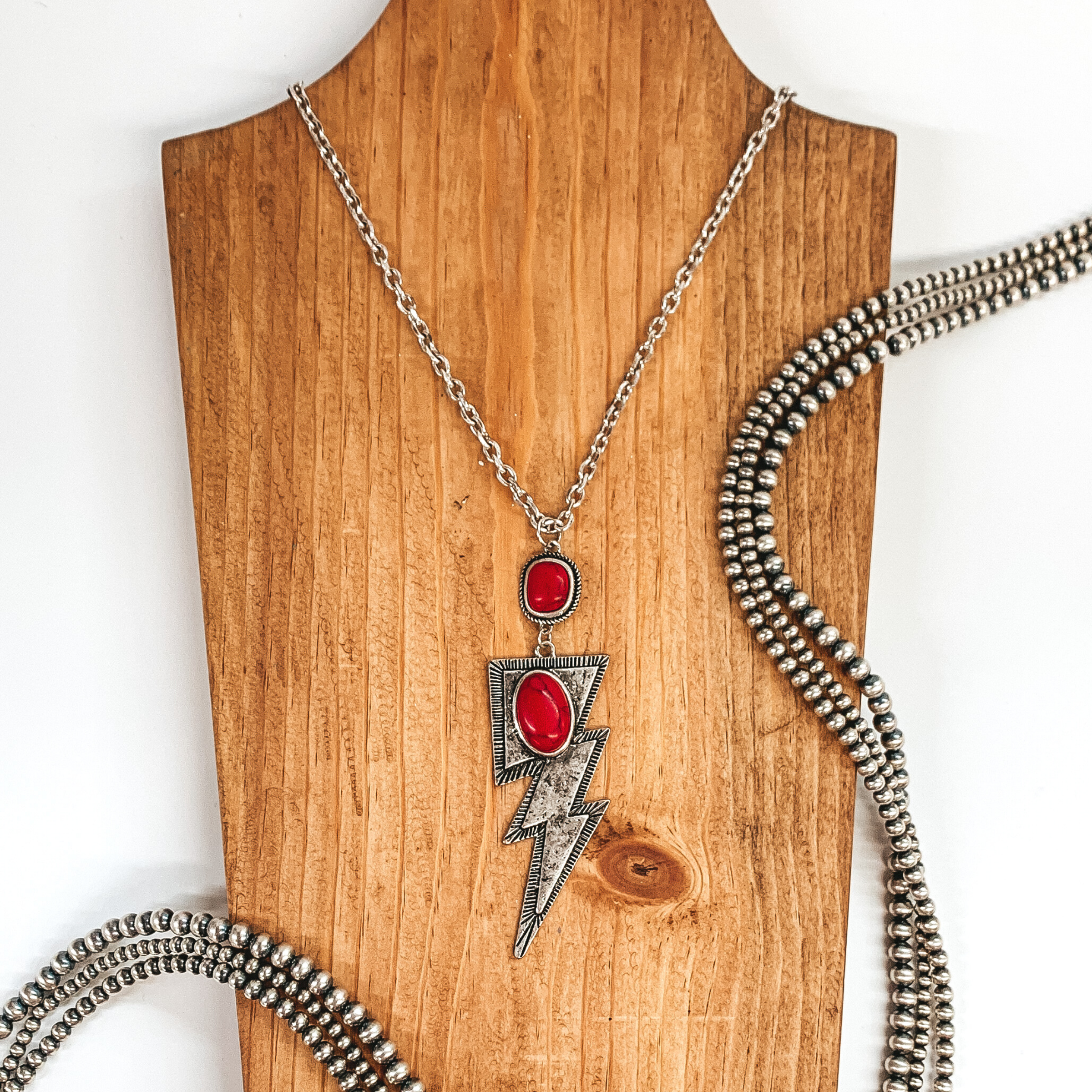 Silver chained necklace with hanging square pendant with red stone and hanging silver lightning bolt pendant with oval red colored stone. This necklace is pictured laying on a tan necklace holder on a white background. This picture includes silver beads as decoration. 