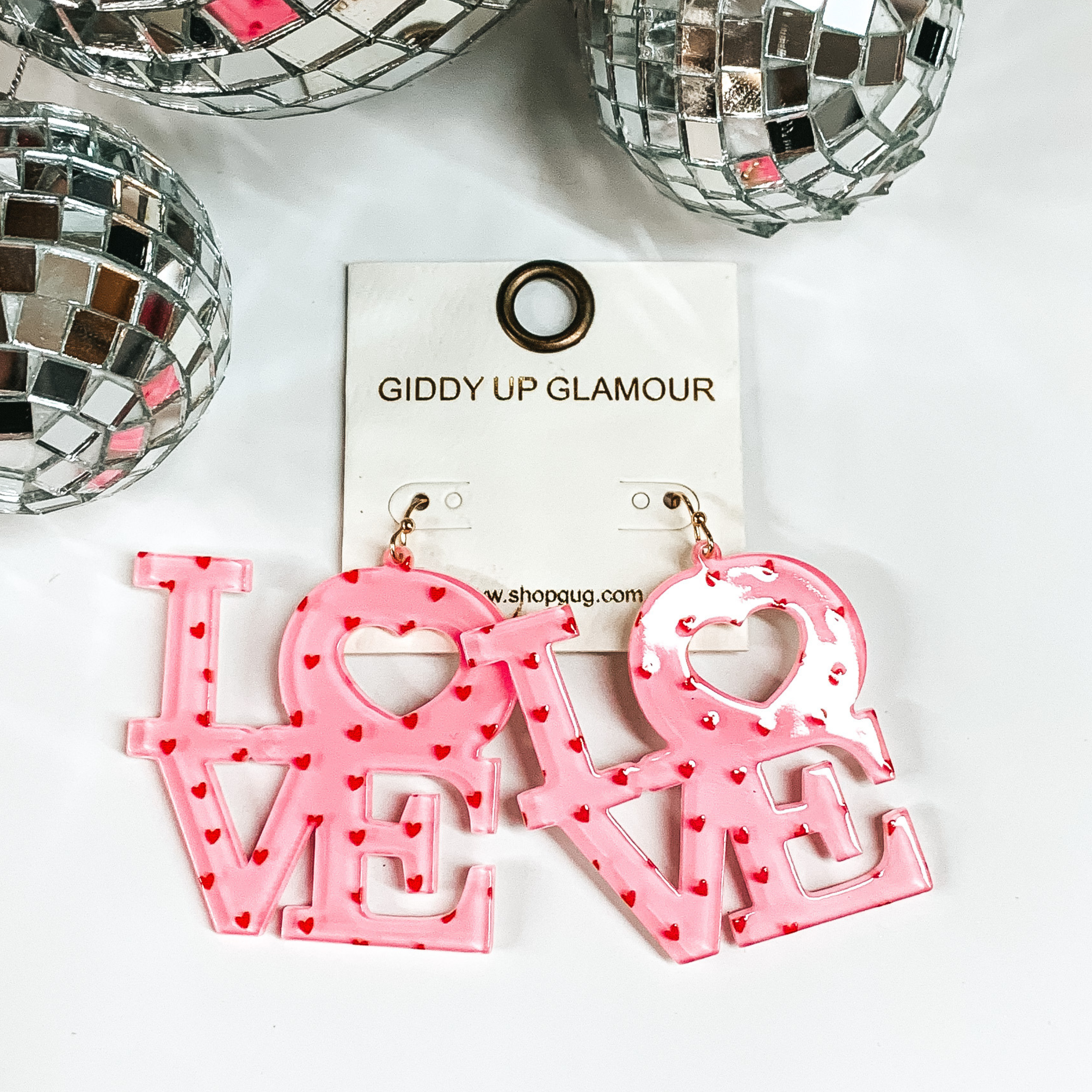 Block letter earrings in light pink that spell out the words LOVE. There are little red hearts on the earrings that add a little extra valentine fun. Earrings are on white back ground with silver disco balls.