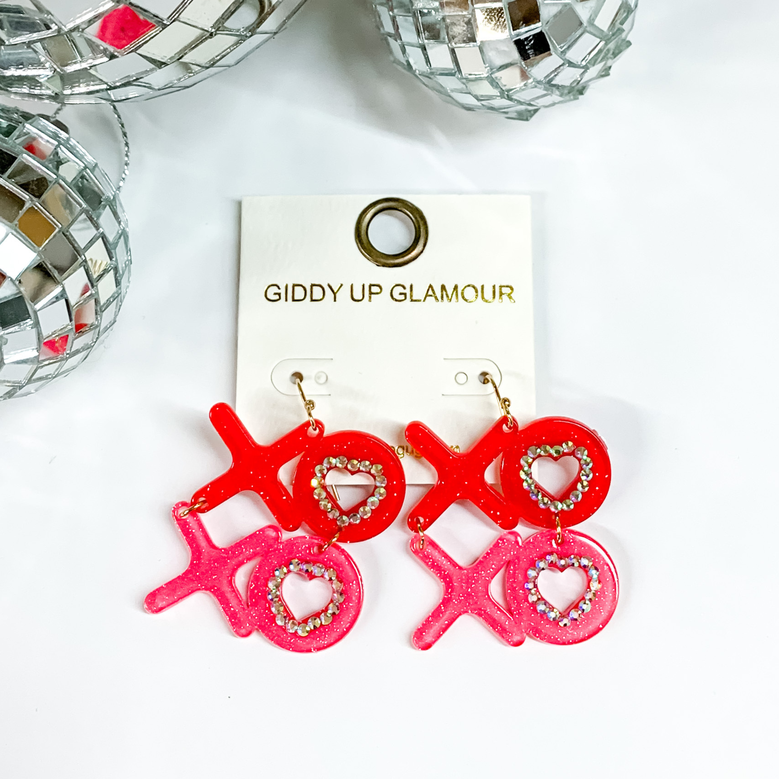 Festive Valentines Day XOXO block letter earrings in red and pink with crystals around the O in XO. Earrings are placed on a white background with silver disco balls  at the top. 