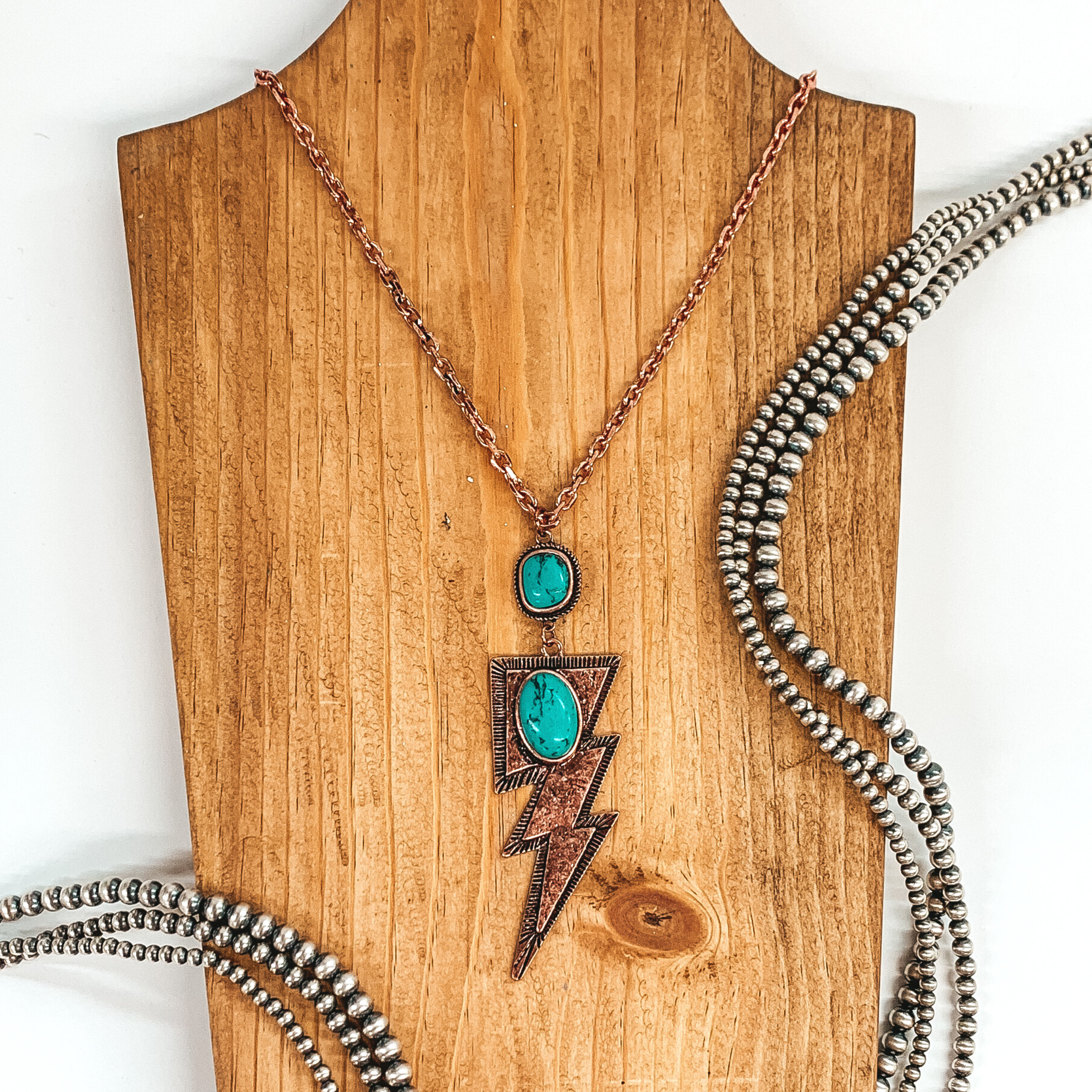 Copper chained necklace with hanging square pendant with turquoise stone and hanging copper lightning bolt pendant with oval turquoise colored stone. This necklace is pictured laying on a tan necklace holder on a white background. This picture includes silver beads as decoration. 