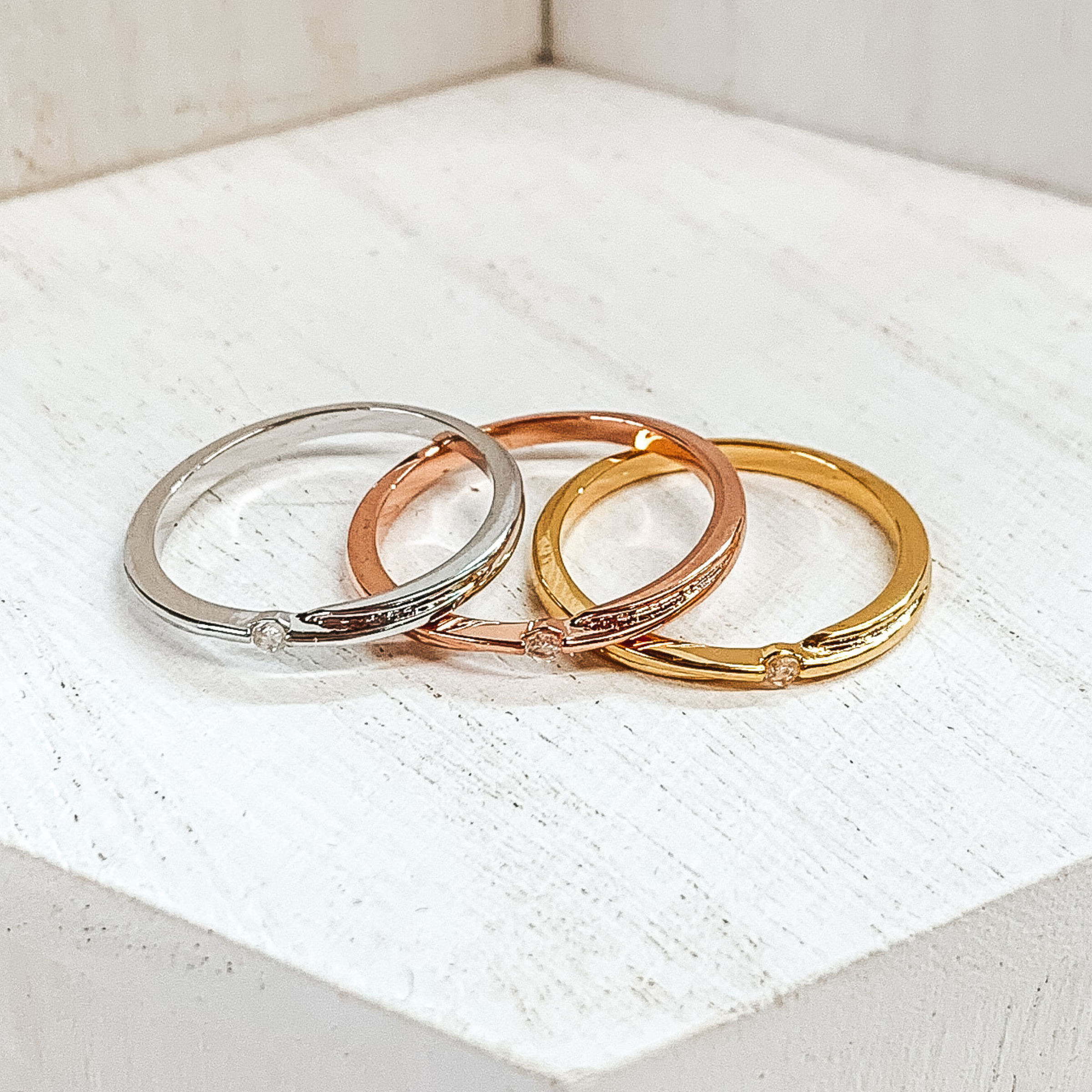Set of three rings in silver, gold, and rose gold. Each ring has a center crystal in the center matching the color of the ring. These rings are pictured on a white background.