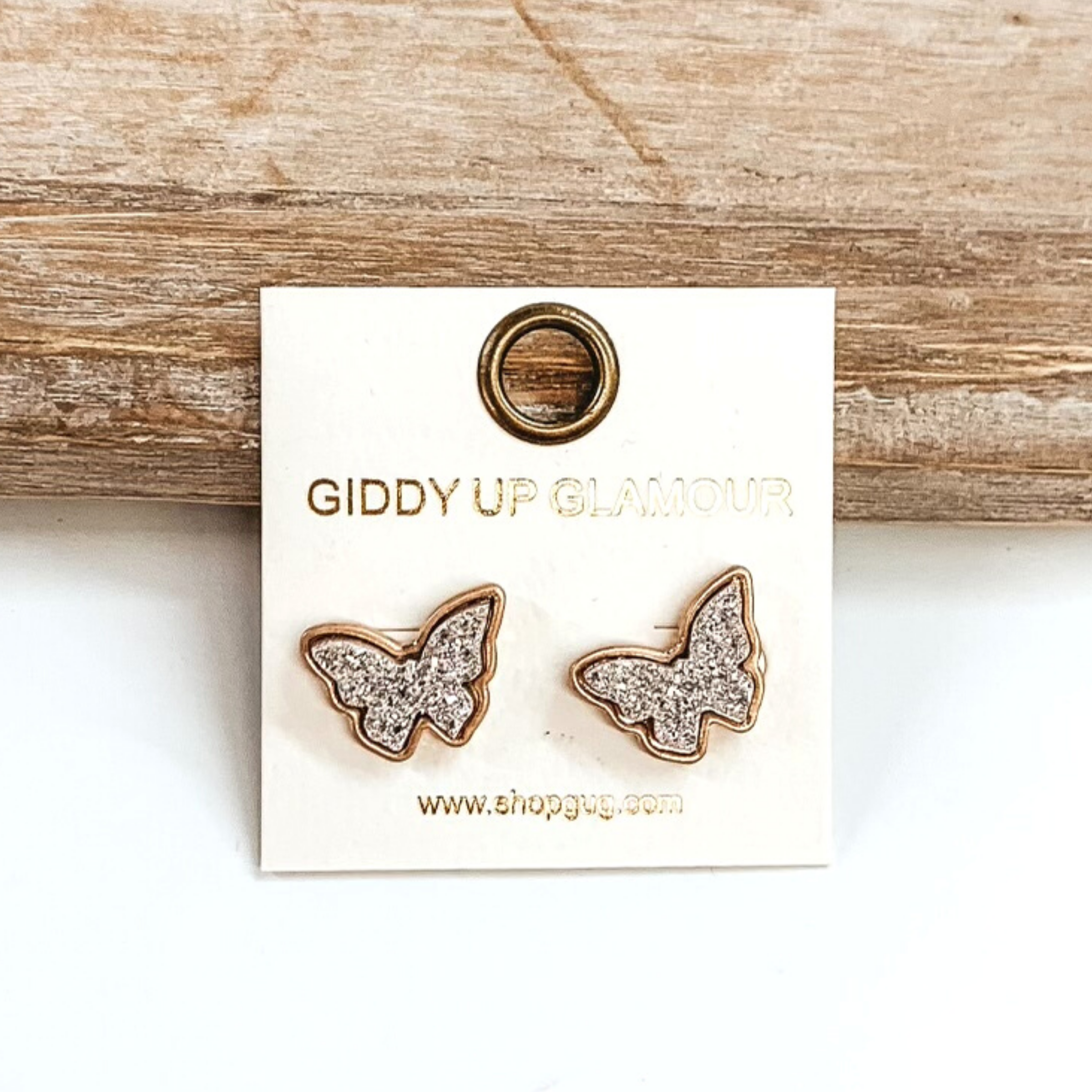 Gold post back earrings with druzy, rose gold, butterfly shaped stone. These earrings are pictured on a white earrings holder on a white background, leaning against a tan block.