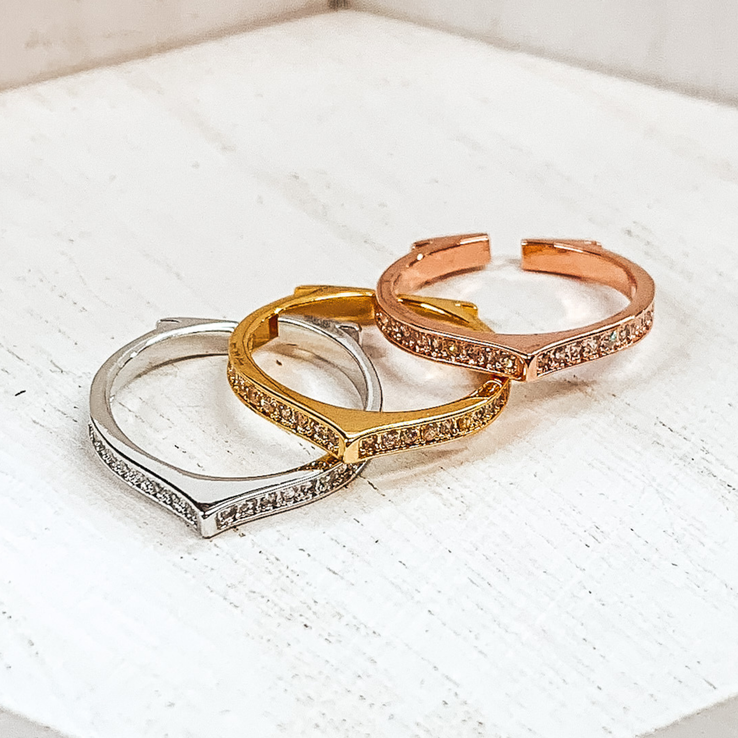 Set of three open rings in silver, gold, and rose gold. Each ring has a pointed center with crystals in the center matching the color of the ring. These rings are pictured on a white background. 