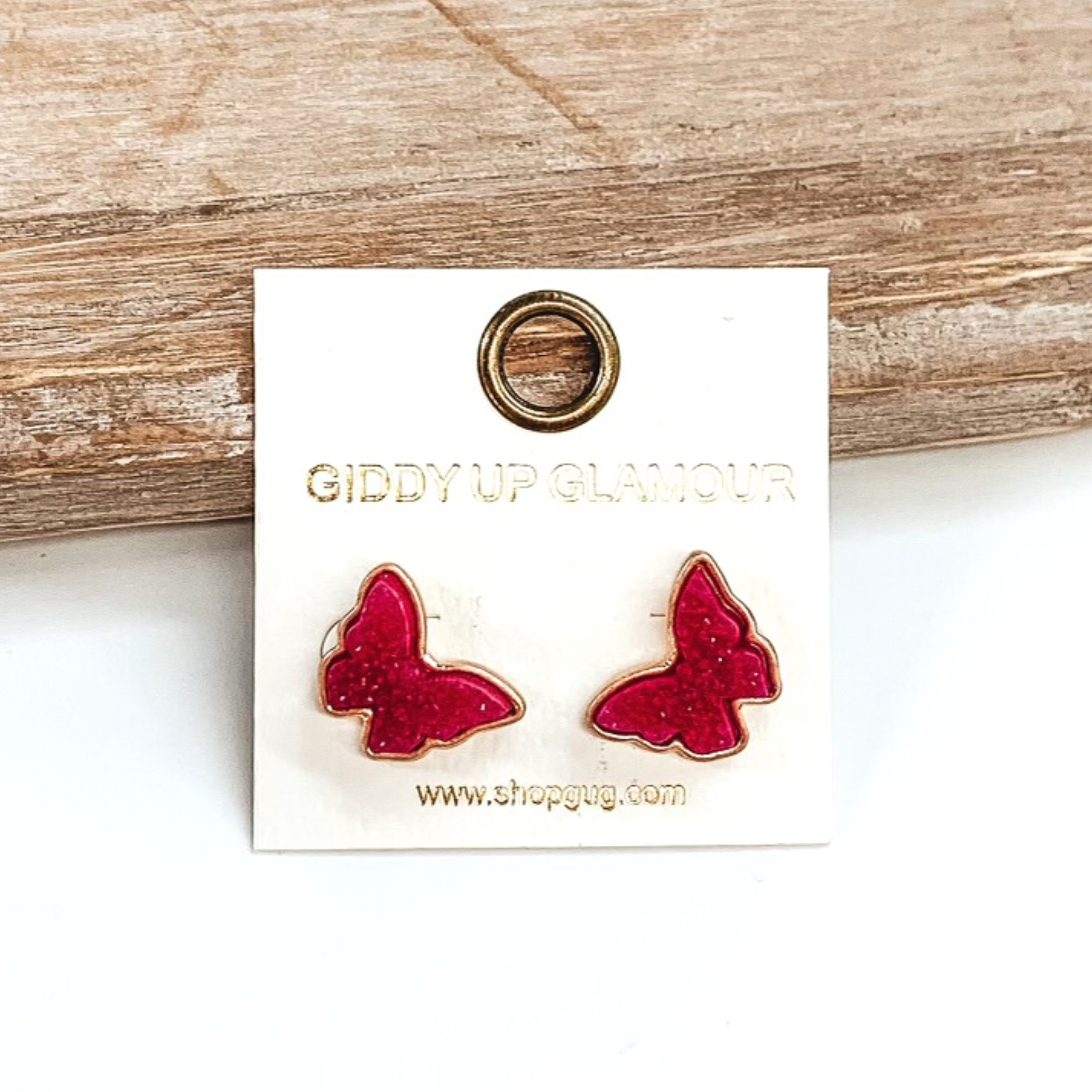 Gold post back earrings with druzy, fuchsia, butterfly shaped stone. These earrings are pictured on a white earrings holder on a white background, leaning against a tan block.