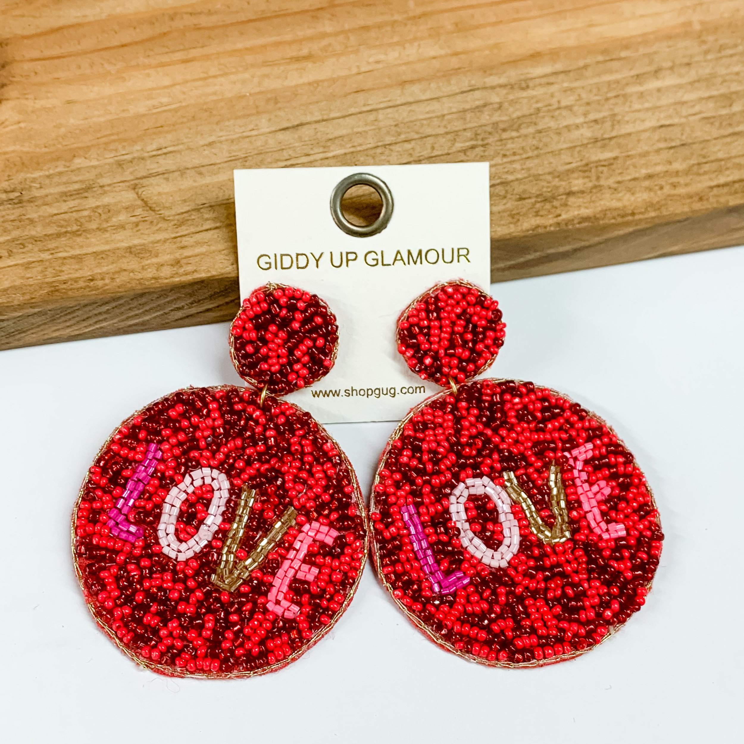 These seedbead earrings have a red mix background with lettering LOVE in the middle of the circle. These red LOVE earrings are perfect for Valentine's Day. These earrings are placed on a white and wood background. 