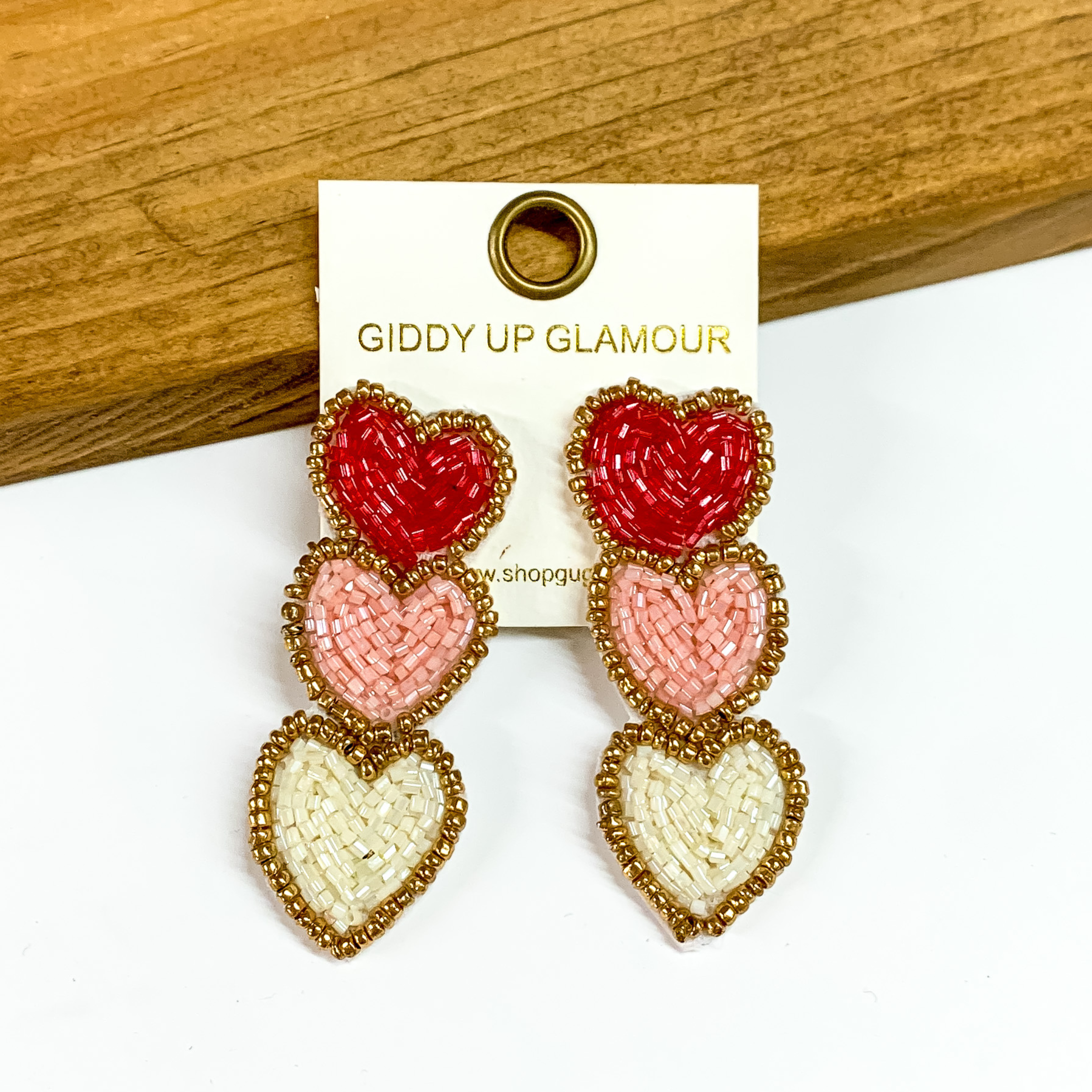 The cutest heart seedbead earrings with post backs. The earrings feature three hearts in different color. The first heart is red, the second heart is light pink and the heart on the bottom is ivory. The earrings are perfect for Valentine's Day. The earrings are placed on a white and wood background.