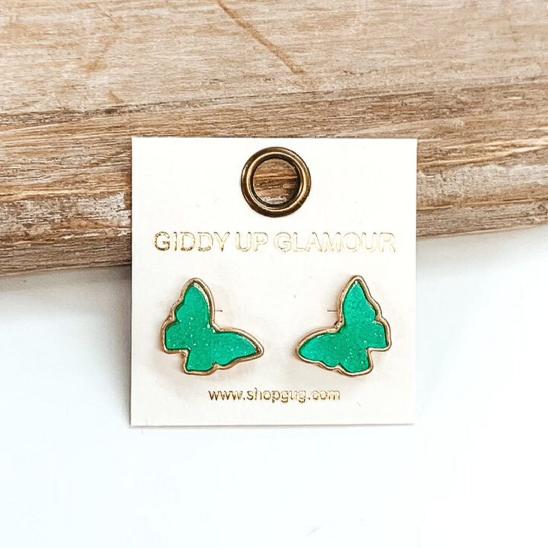 Gold post back earrings with druzy, mint, butterfly shaped stone. These earrings are pictured on a white earrings holder on a white background, leaning against a tan block.