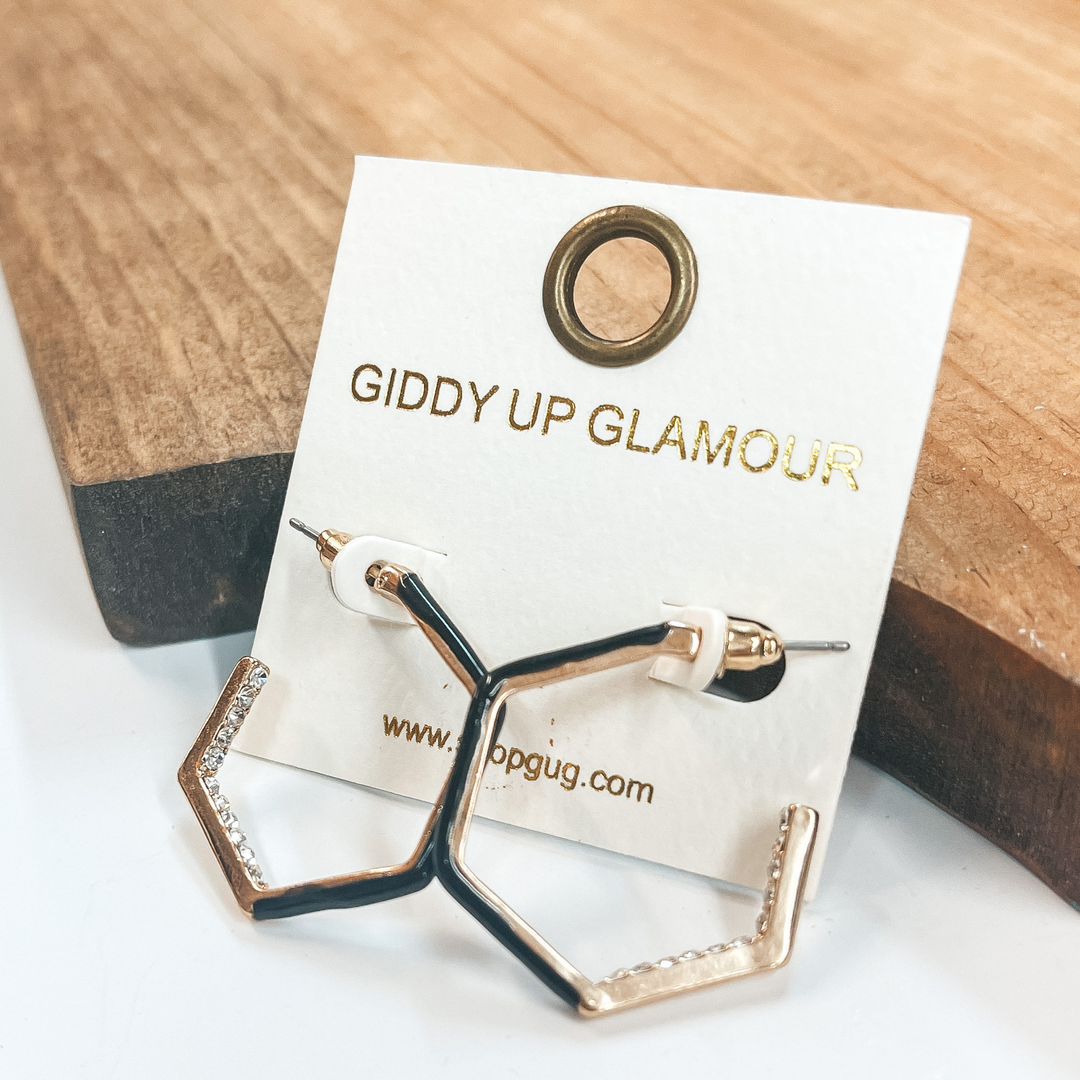 Open ended hexagon hoops in gold  with clear crystals on the inside  of two sides and black on the other three sides.  Taken on a white background and leaned up against  a brown block.