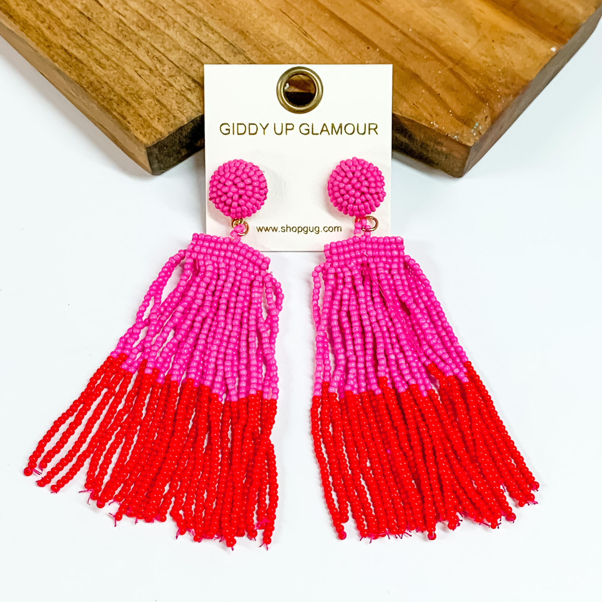 These two toned beaded earrings feature a post back circle with a fringe tassel in seedbeads. The beads start out hot pink and turn to red color block. The earrings are laying on a white and wood background. 