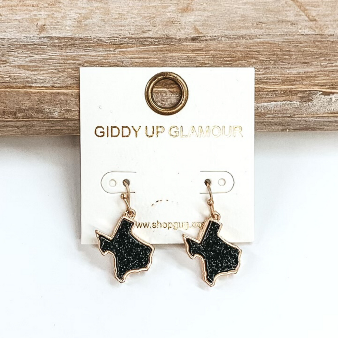 Gold dangle earrings with druzy, black, texas shaped stone. These earrings are pictured on a white earrings holder on a white background, leaning against a tan block.
