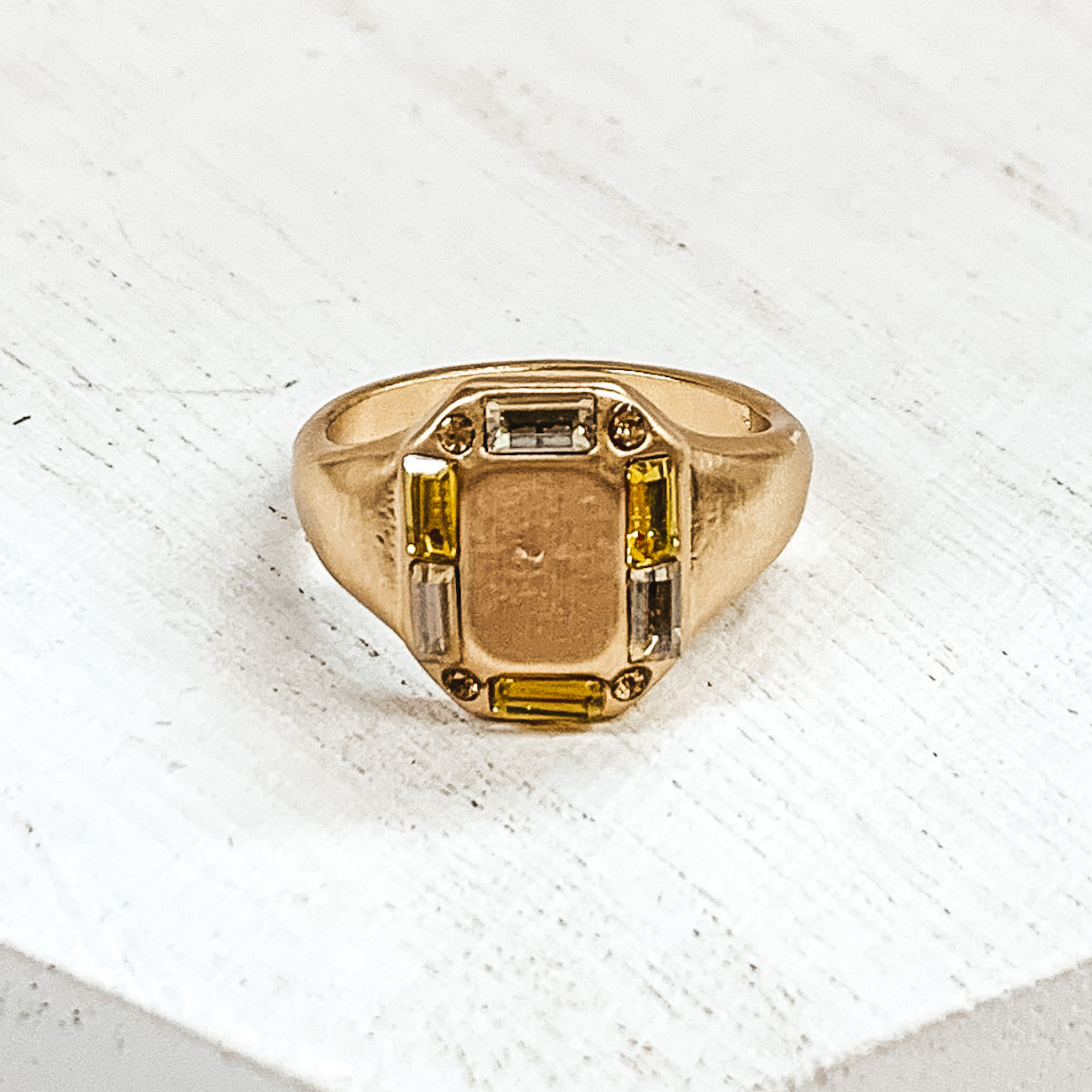 Thick gold band with a rectangle shaped front. The rectangle part includes small, mix of yellow crystals outlining it. This ring is pictured on a white background.