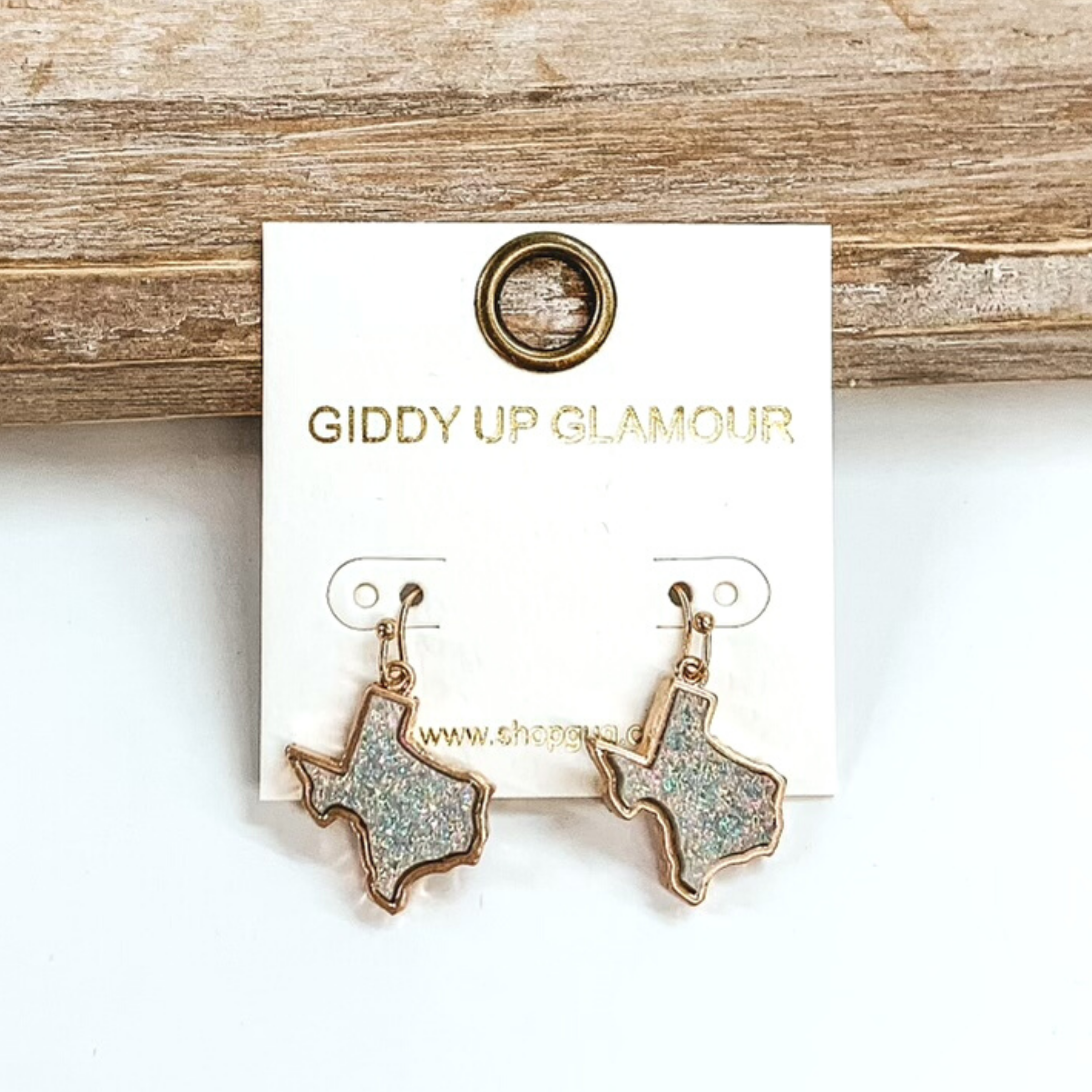 Gold dangle earrings with druzy, grey iridescent, texas shaped stone. These earrings are pictured on a white earrings holder on a white background, leaning against a tan block.