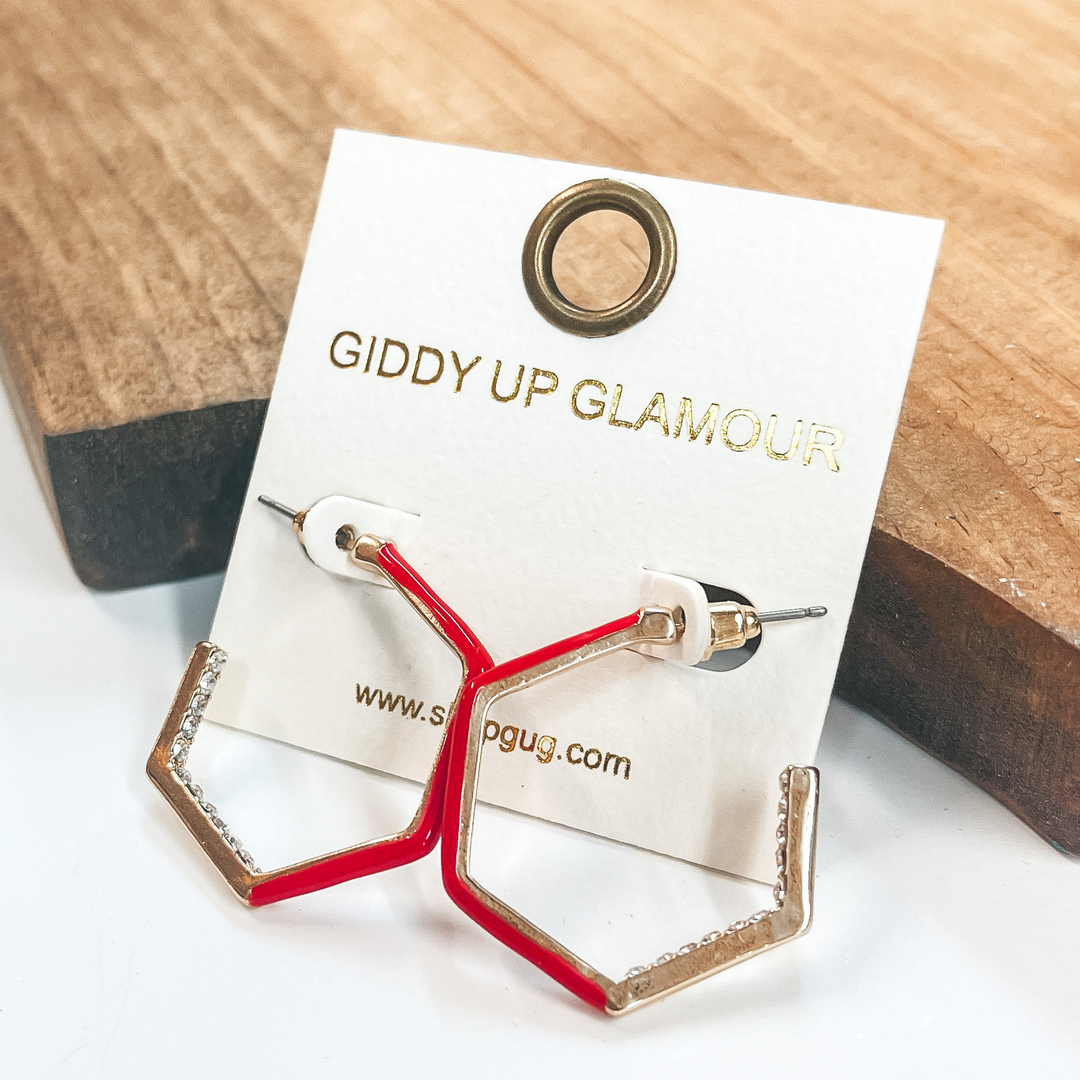 Open ended hexagon hoops in gold  with clear crystals on the inside  of two sides and red on the other three sides.  Taken on a white background and leaned up against  a brown block.
