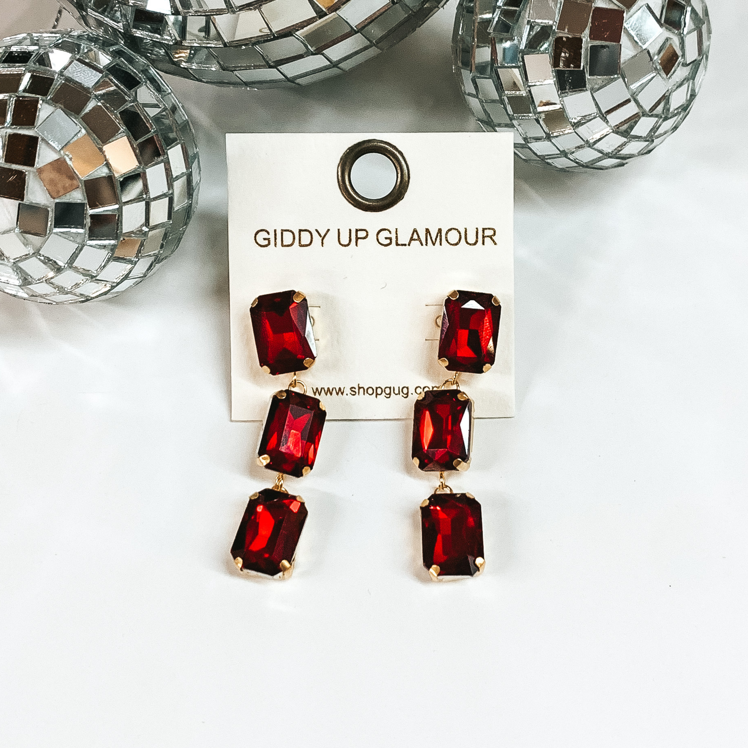 These stunning earrings add the perfect amount of sparkle. Earrings feature three red rectangle crystal stones in a gold tone setting. Earrings are placed on a white board with three disco balls at the top.