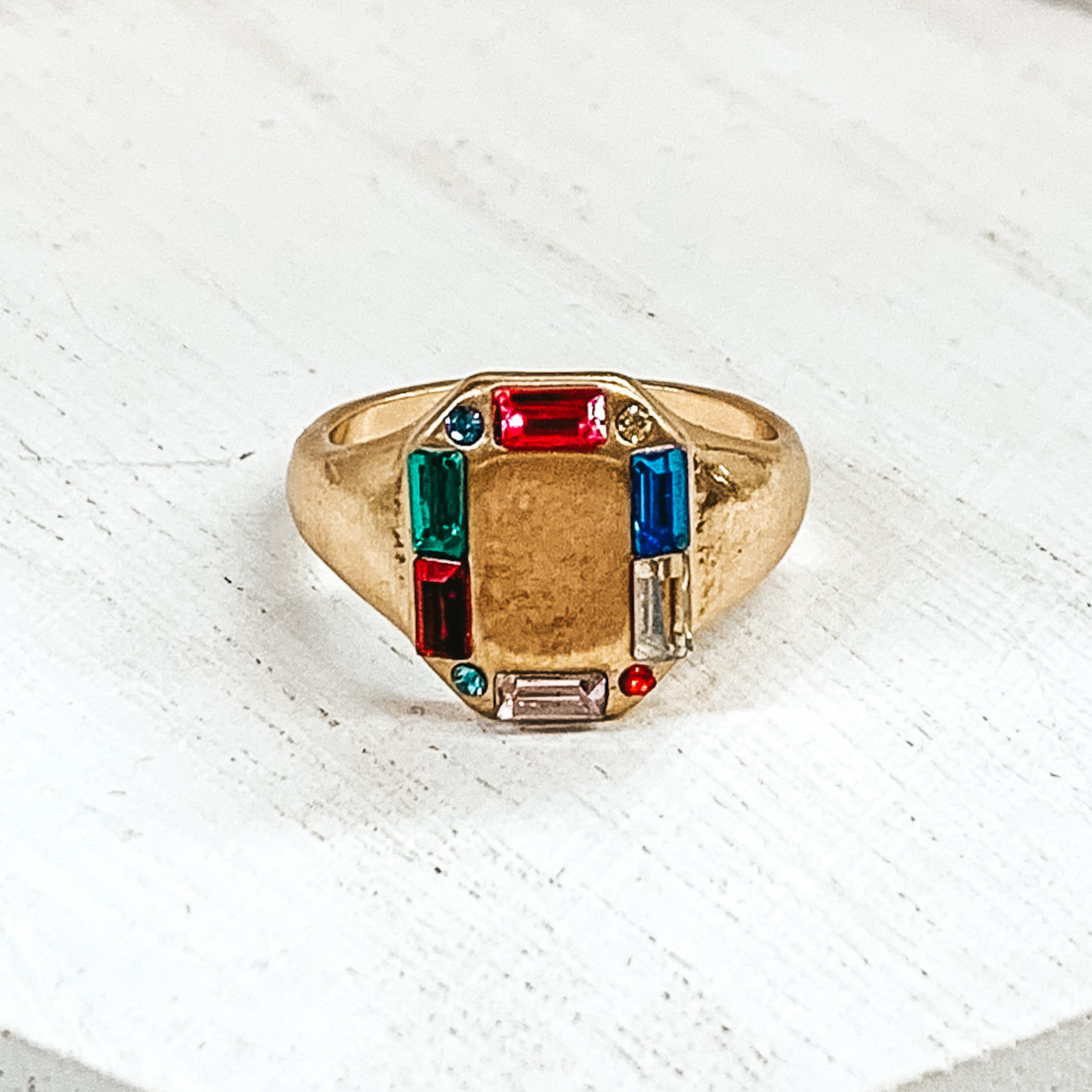 Thick gold band with a rectangle shaped front. The rectangle part includes small, multicolored crystals outlining it. This ring is pictured on a white background. 