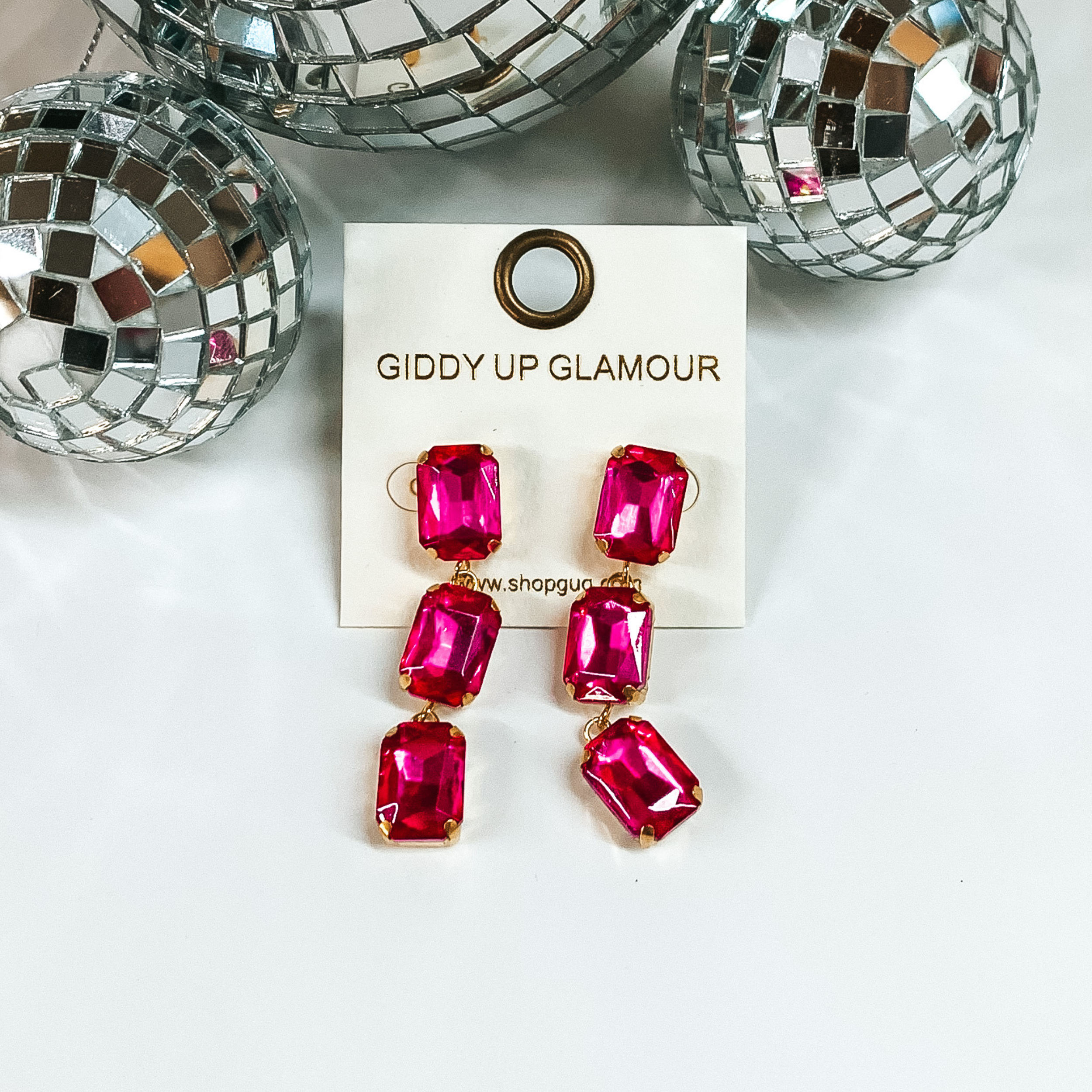 These stunning earrings add the perfect amount of sparkle. Earrings feature three rectangle crystal stones in a gold tone setting. Earrings are placed on a white board with three disco balls at the top. 