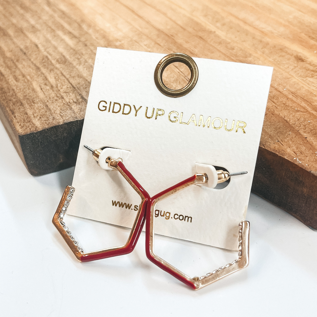 Open ended hexagon hoops in gold  with clear crystals on the inside  of two sides and burgundy on the other three sides.  Taken on a white background and leaned up against  a brown block.