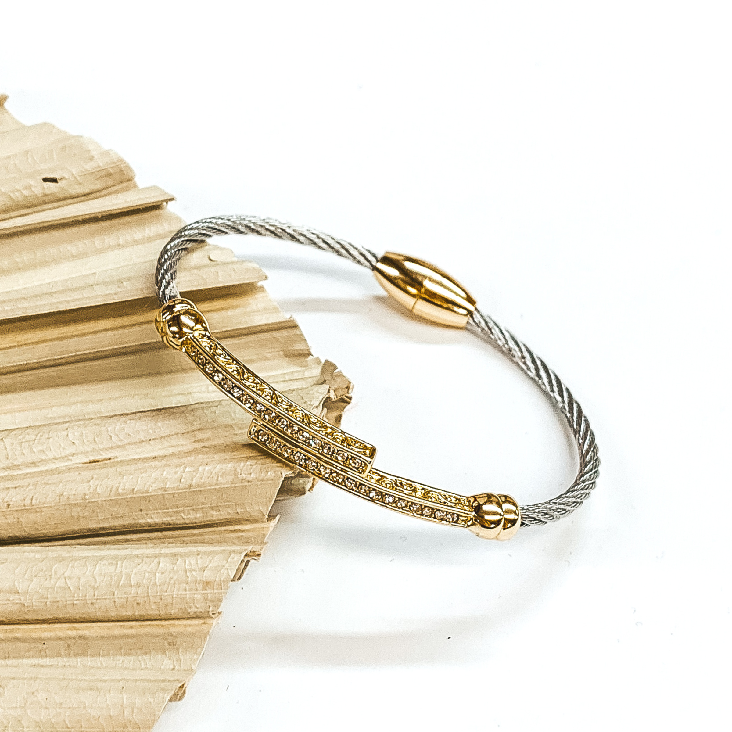 This is a silver cable bracelet with a gold magnetic clasp. It also includes two gold bar attached in the middle with clear crystals. This bracelet is pictured laying on a green palm leaf on a white background. 