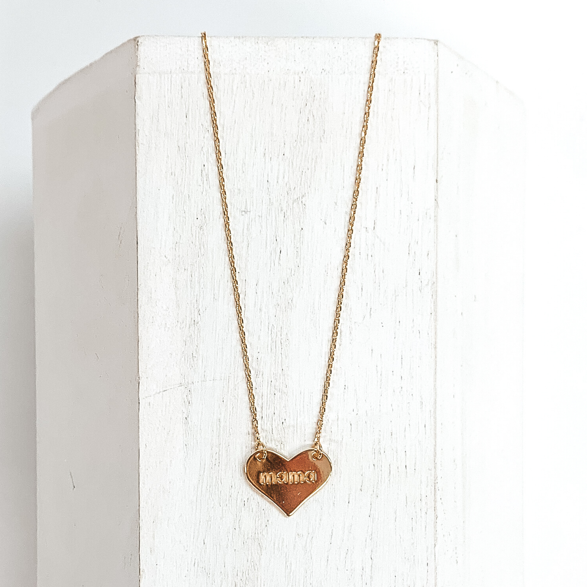 Dainty gold chained necklace with a gold heart pendant. In the center of the pendant has the word "mama" engraved. This necklace is pictured laying on a white block on a white background.