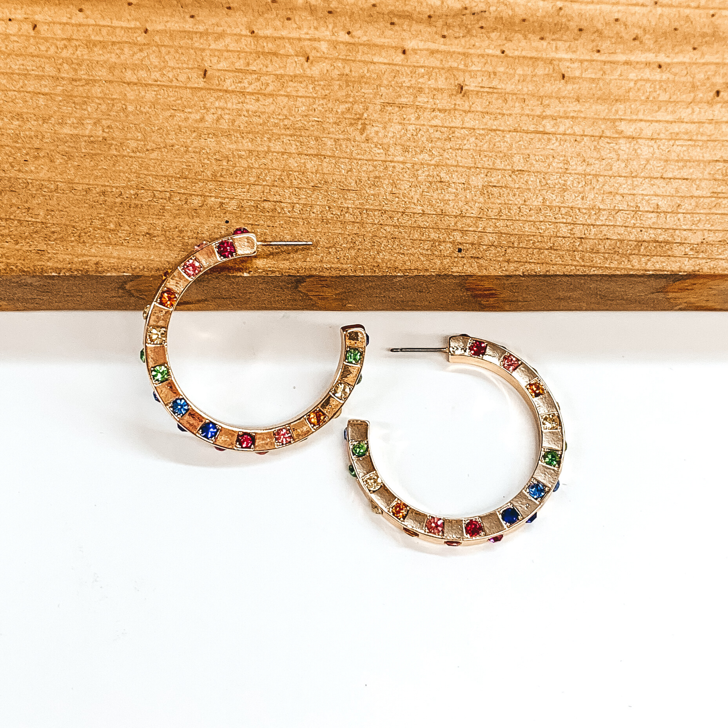 Gold hoop earrings with spaced out multicolored crystals. One earrings is pictured laying on a brown block while the other earrings is pictured laying flat on the white background.