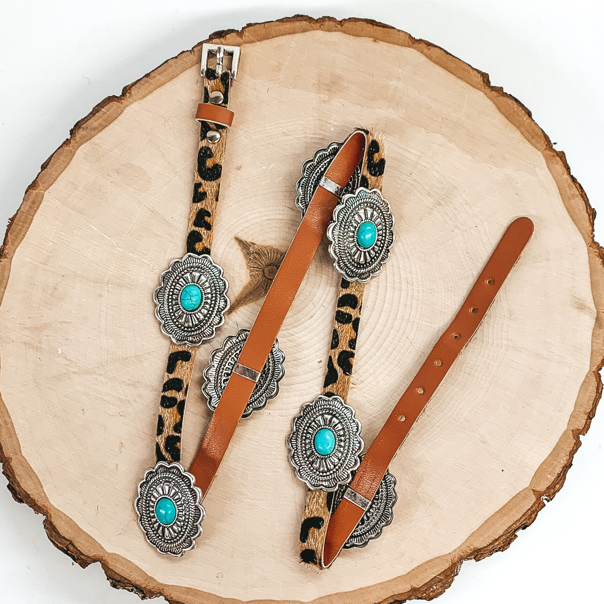 Leopard print hat band with silver, spaced out conchos. The conchos has turquoise center stones. This hat band is pictured laying on a piece of wood on a white background.