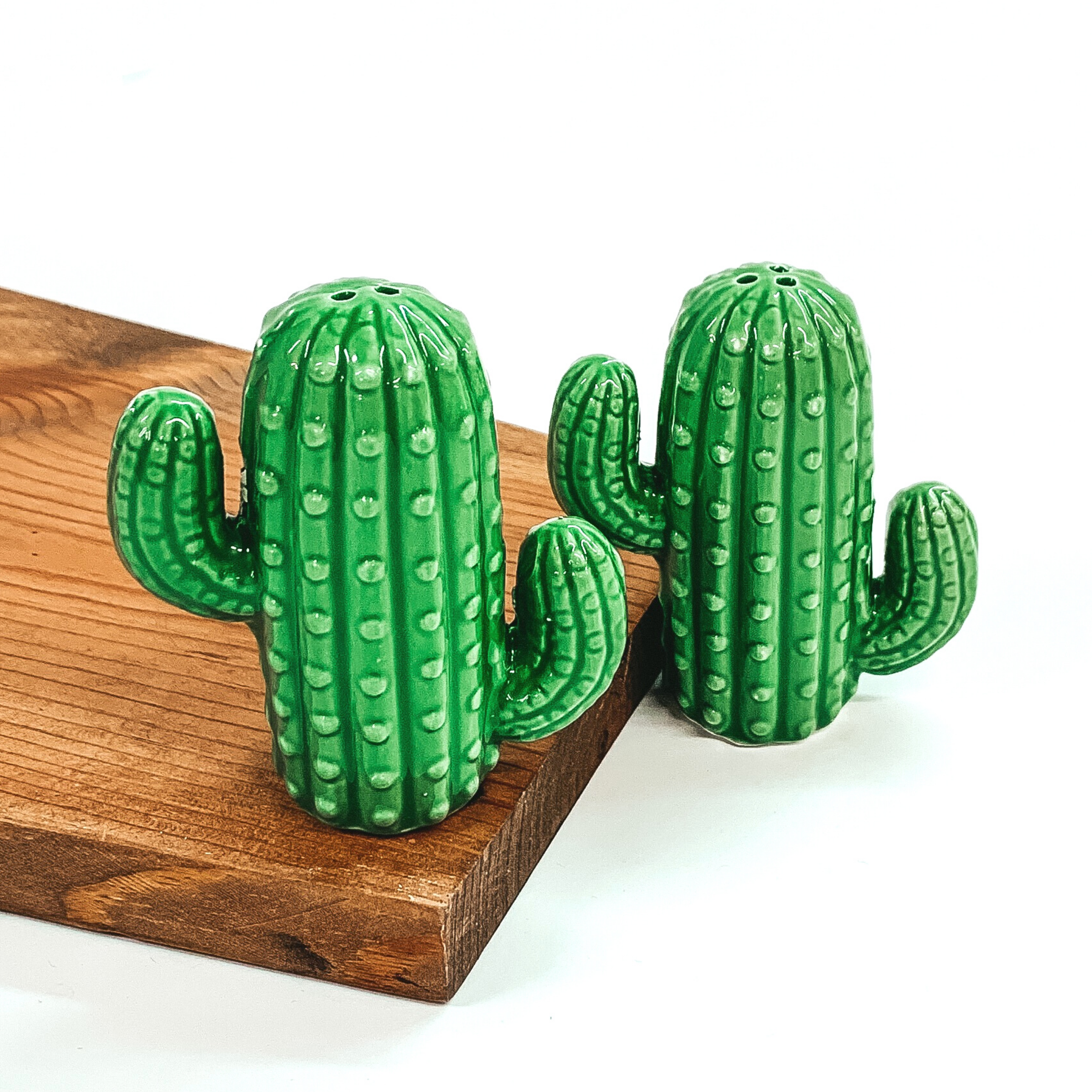 Green cactus salt and pepper shakers. One is on top of a dark piece of wood, white the other is standing next to it. The shakers are pictured on a white  background. 