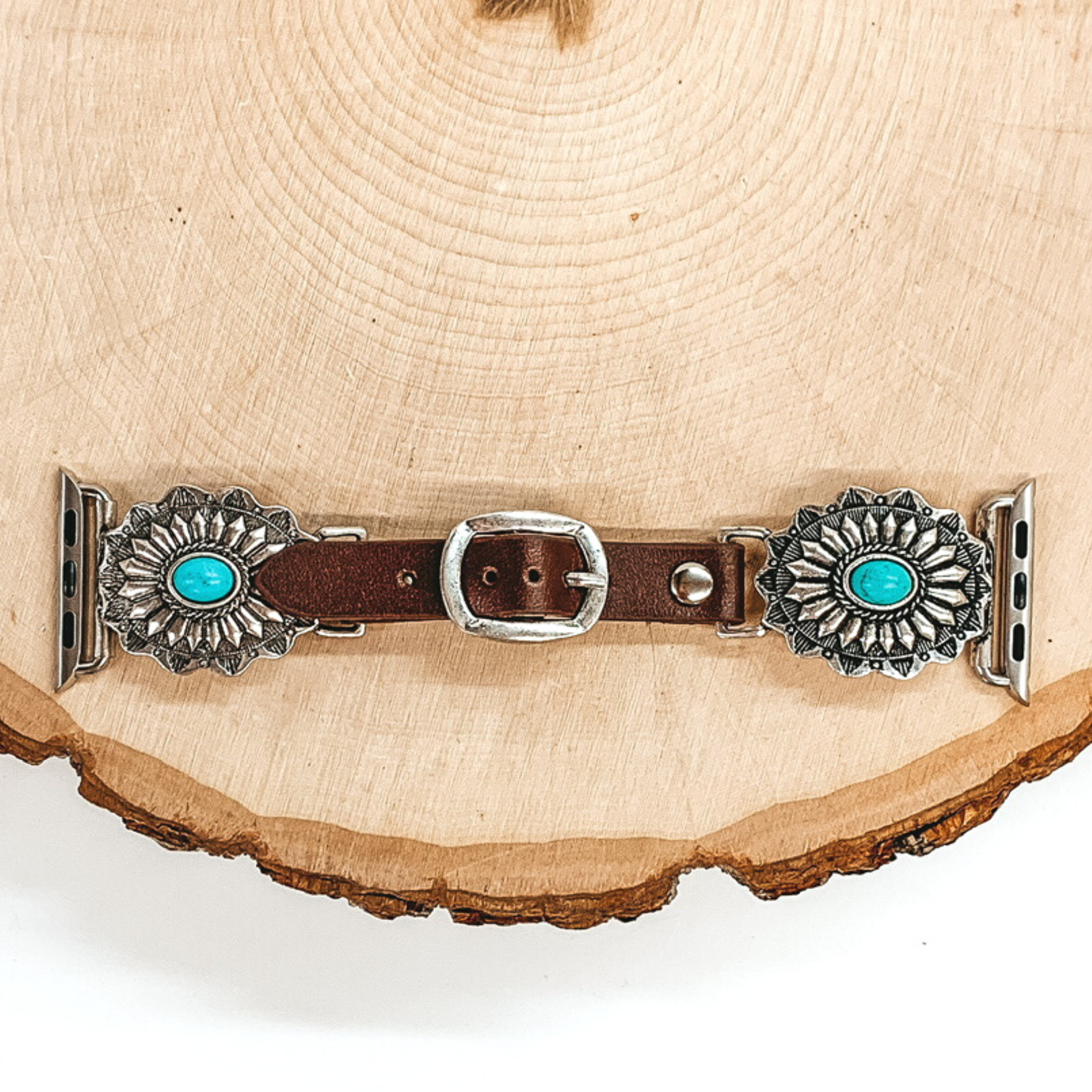 Dark Brown watch band with silver, oval concho ends with Apple watch band acessories. The conchos have small, center turquoise stone. This watch band is pictured on a piece of wood on a white background.