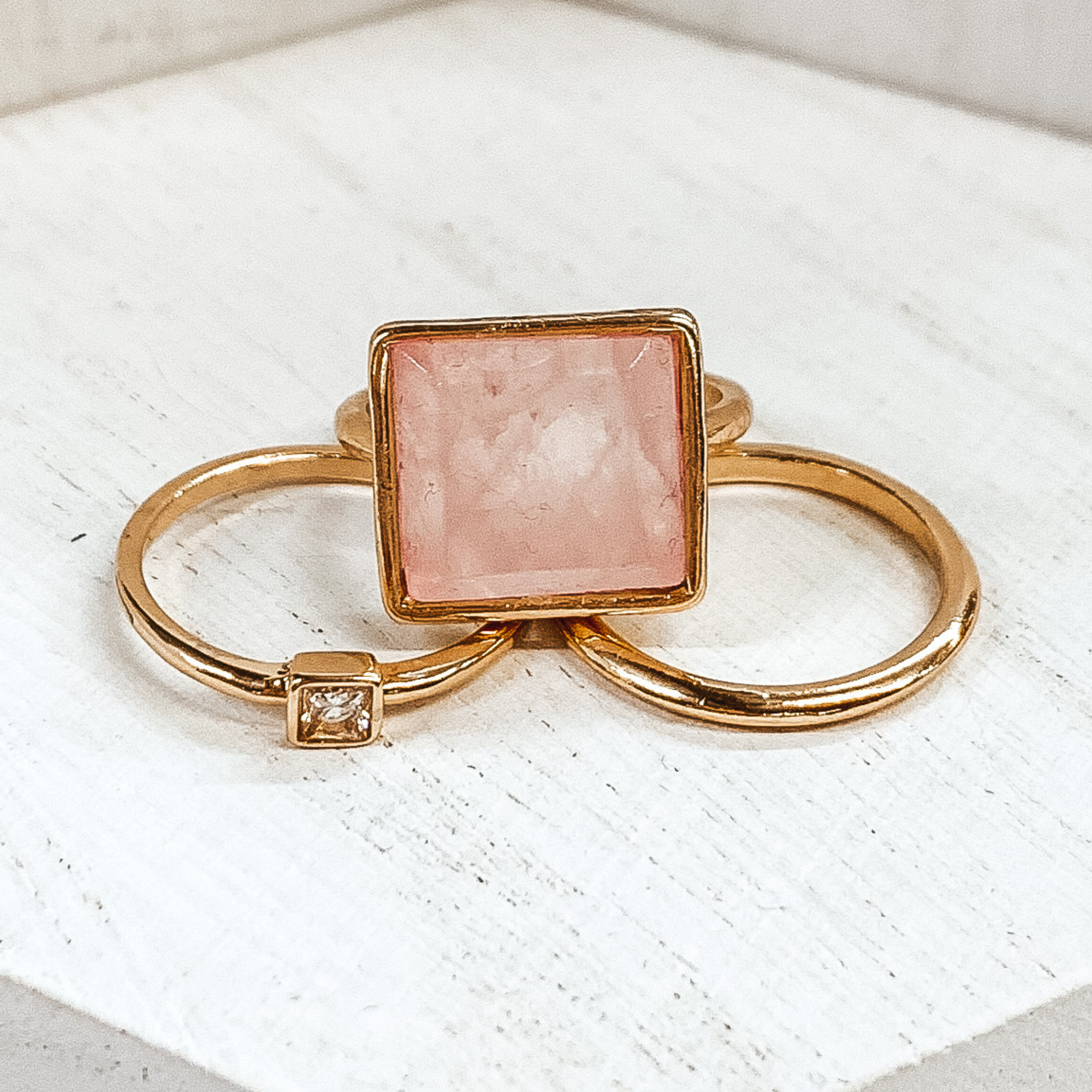 Set of three gold rings. One ring is a plain gold band. Another has a small square pendant with a center crystal. The third ring has a big, light pink pendant. These rings are pictured on a white background.  