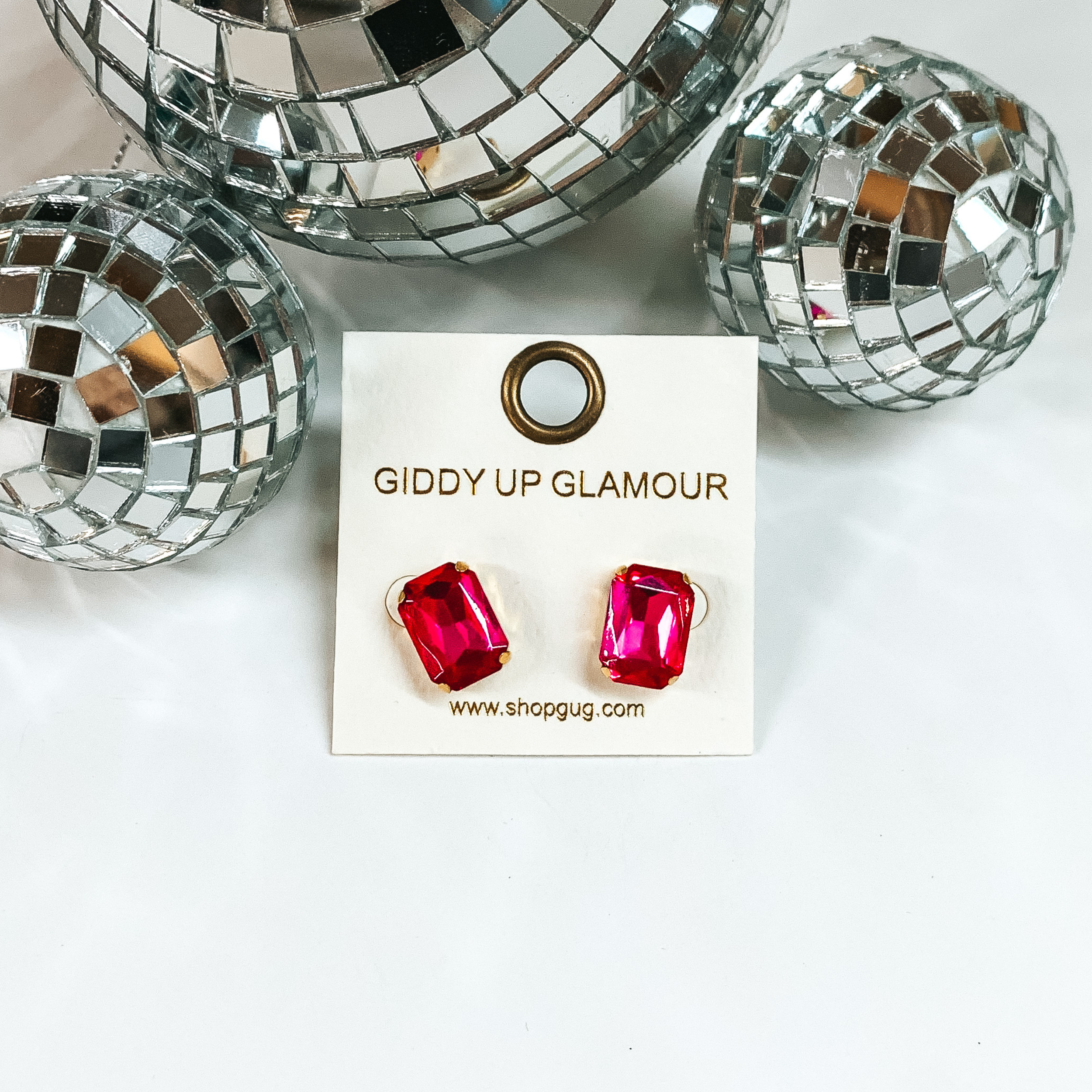 These stunning earrings add the perfect amount of sparkle. Earrings feature a hot pink rectangle crystal stone in a gold tone stud setting. Earrings are placed on a white background with silver disco balls at the top. 