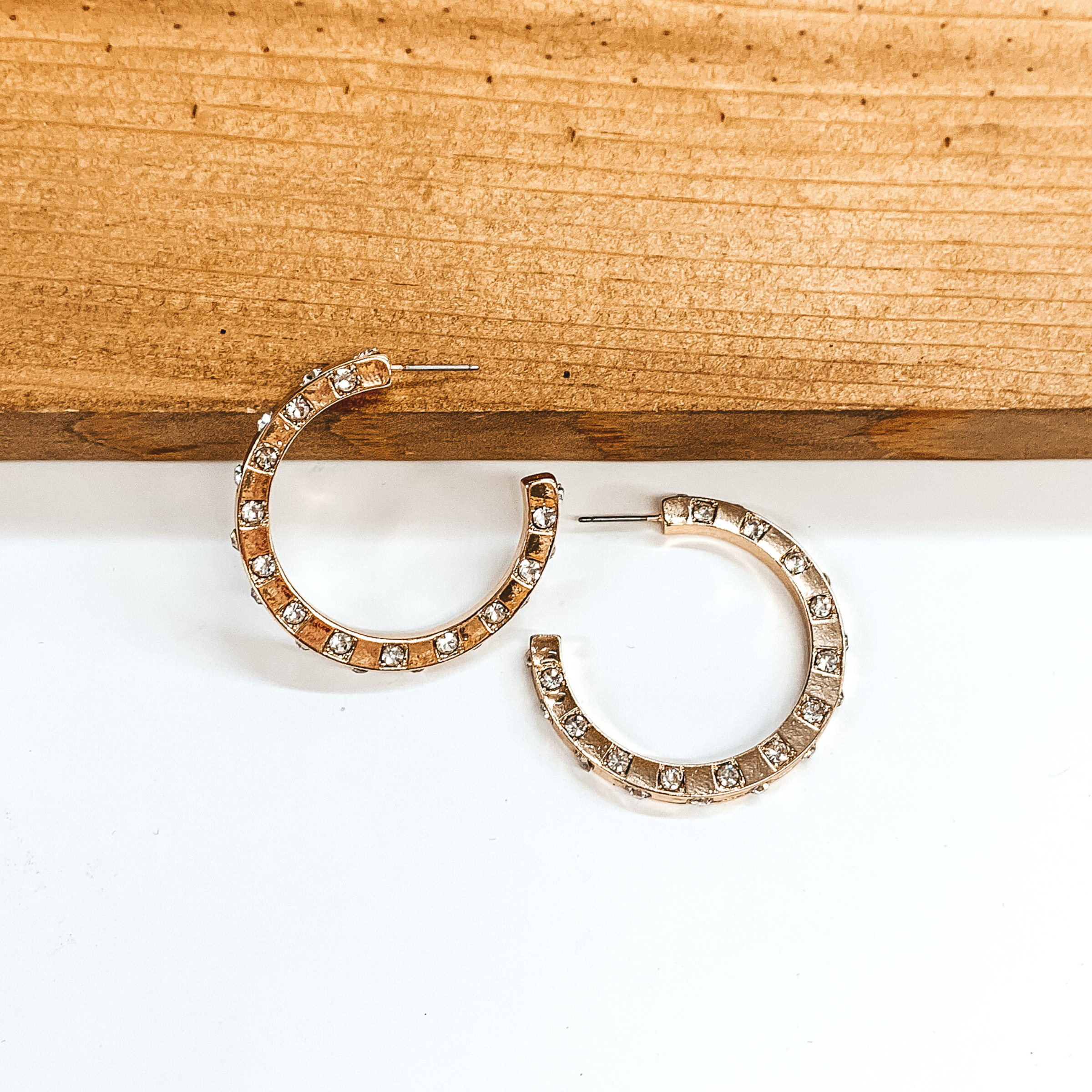 Gold hoop earrings with spaced out clear crystals. One earrings is pictured laying on a brown block while the other earrings is pictured laying flat on the white background.