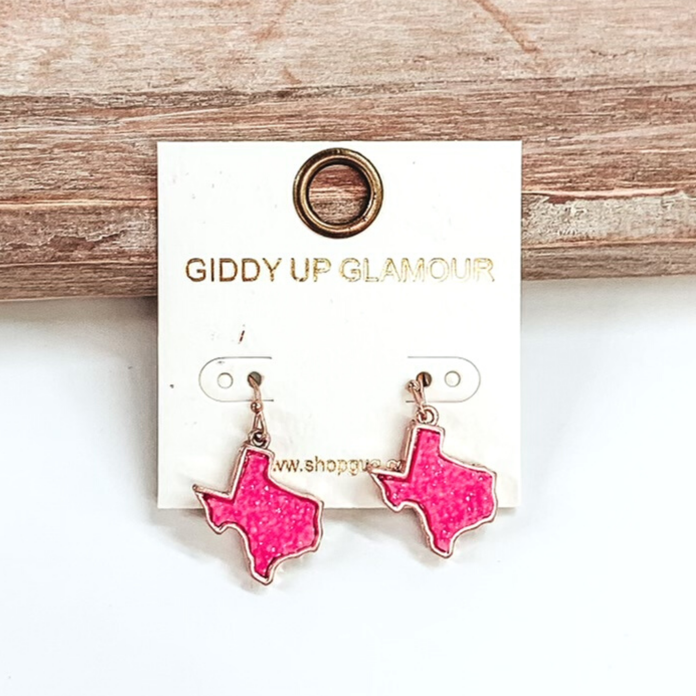 Gold dangle earrings with druzy, neon pink, texas shaped stone. These earrings are pictured on a white earrings holder on a white background, leaning against a tan block.