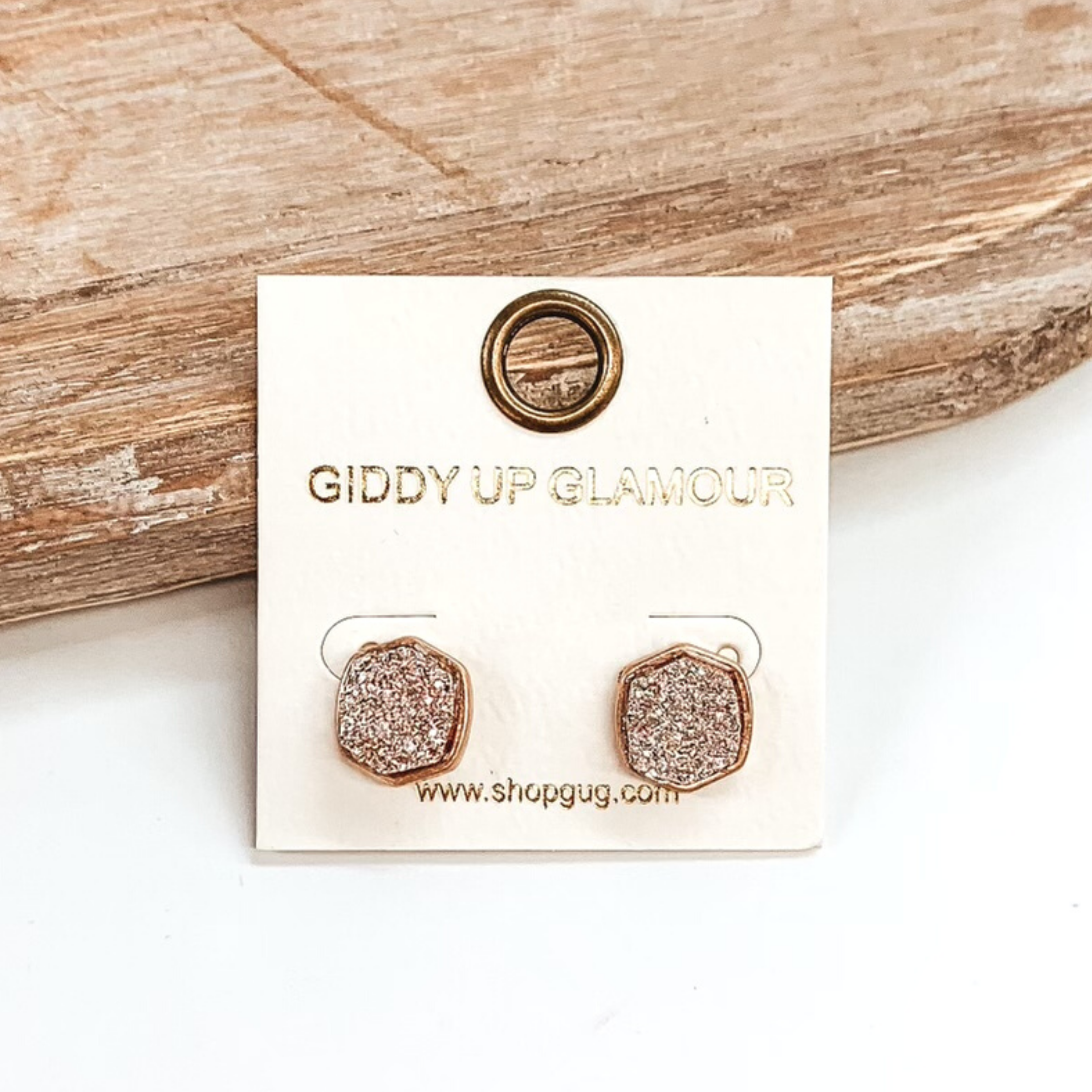 Gold post back earrings with druzy, rose gold, hexagon shaped stone. These earrings are pictured on a white earrings holder on a white background, leaning against a tan block.