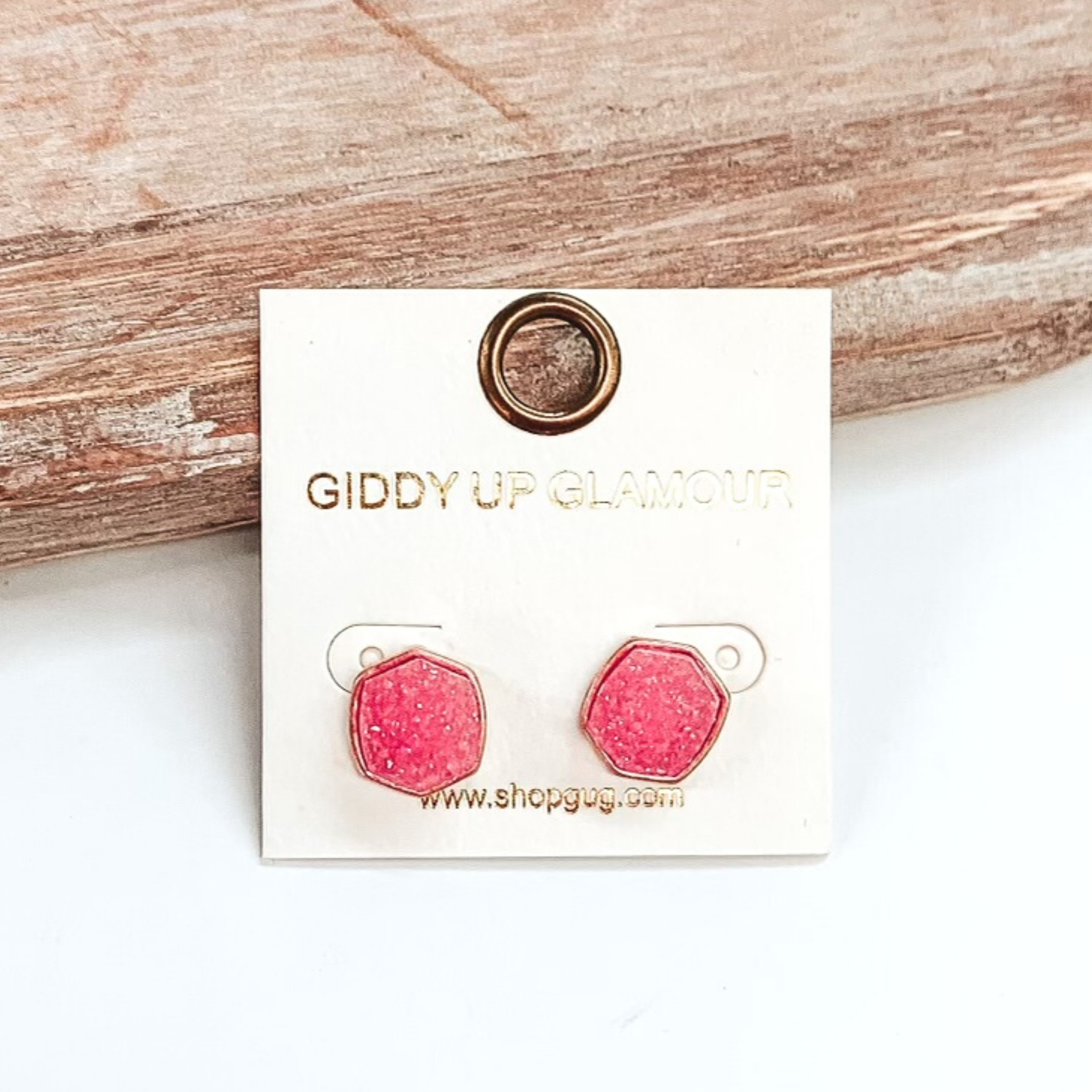 Gold post back earrings with druzy, pink, hexagon shaped stone. These earrings are pictured on a white earrings holder on a white background, leaning against a tan block.