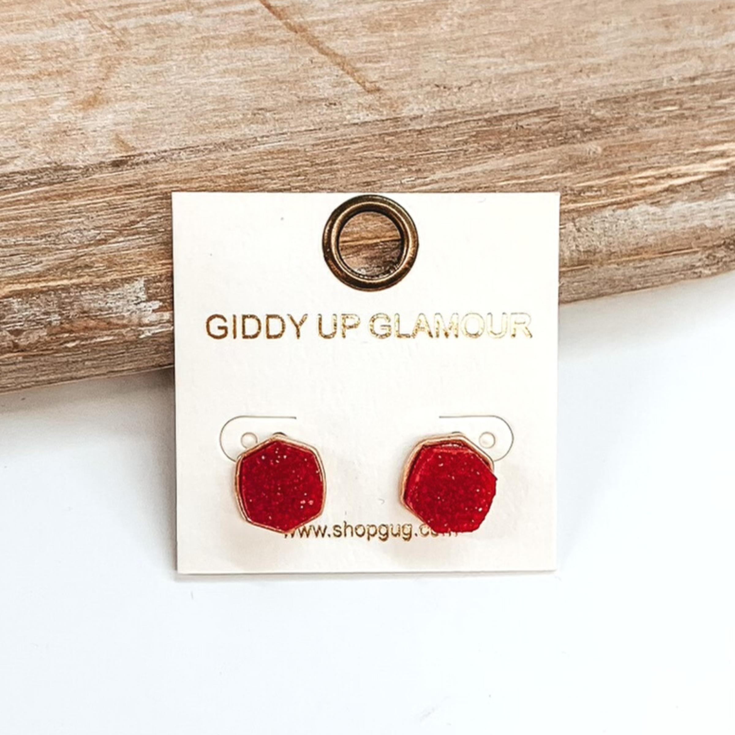Gold post back earrings with druzy, red, hexagon shaped stone. These earrings are pictured on a white earrings holder on a white background, leaning against a tan block.