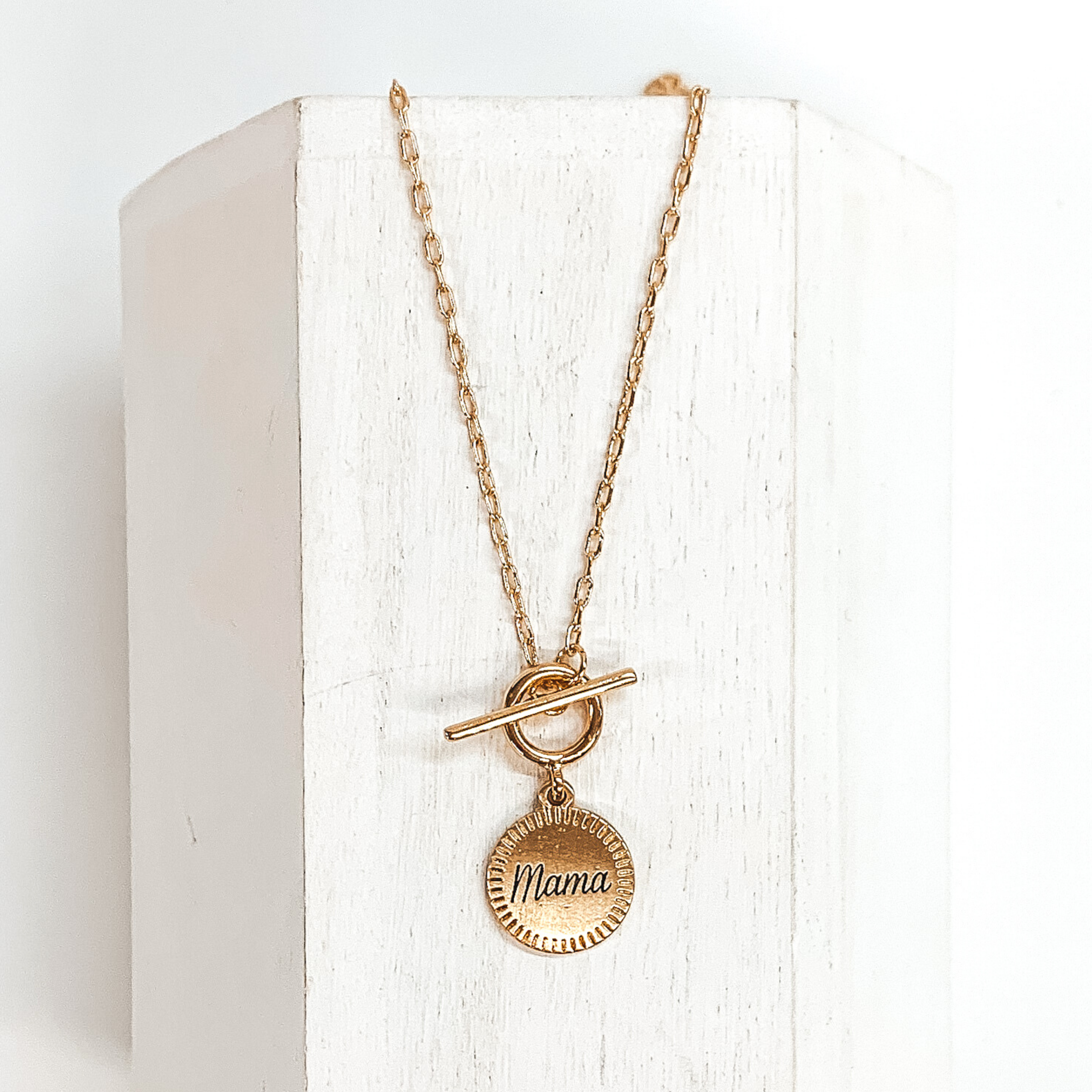 This is a gold chained necklace with a front toggle clasp that has a hanging circle pendant. In the center of the pendant the word "Mama" is engraved in cursive. This necklace is pictured laying on a white block on a white background.