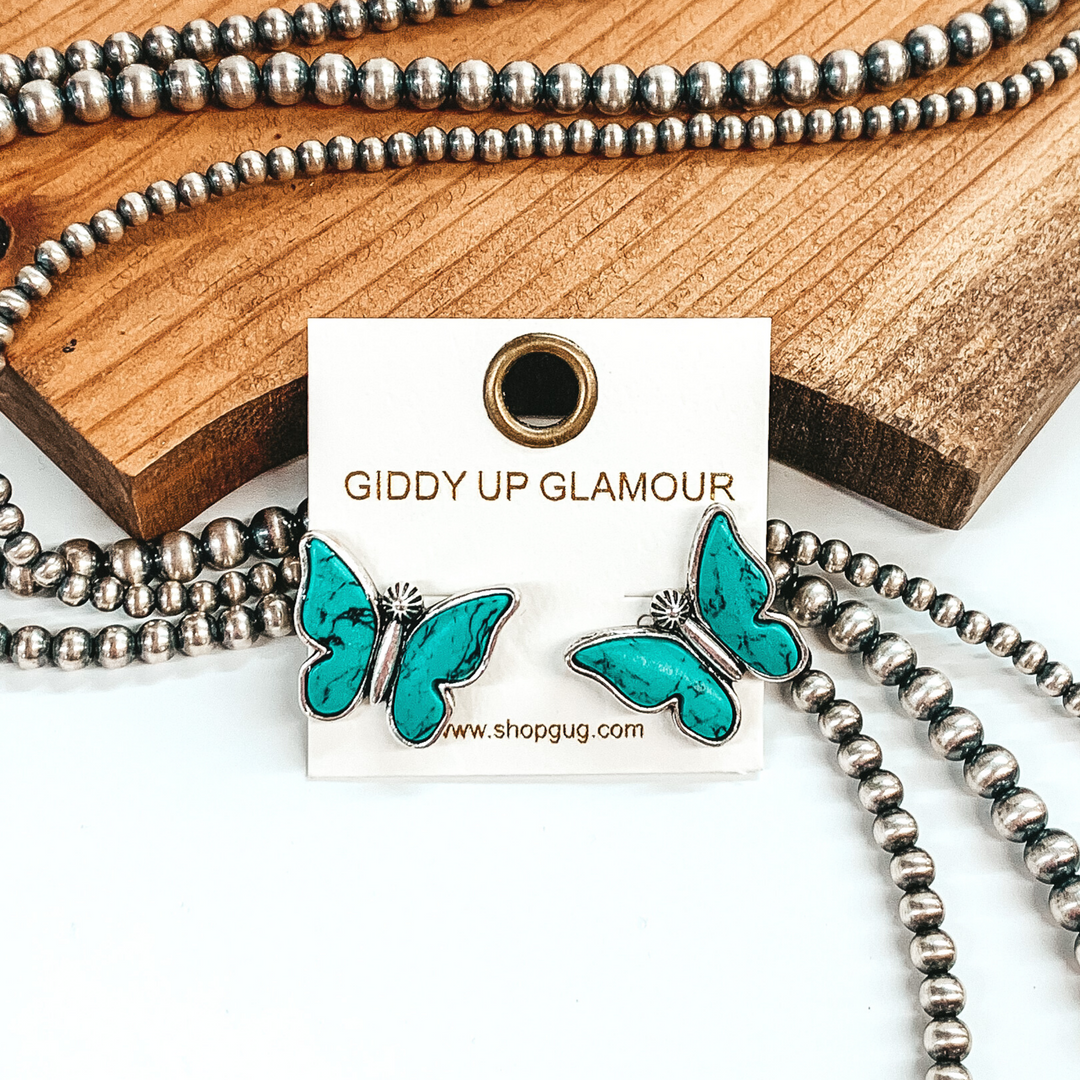 Silver post stud earrings shaped like a butterfly with turquoise stones for wings. These earrings are pictured in front of brown wood with some silver pearls laying behind the earrings. 