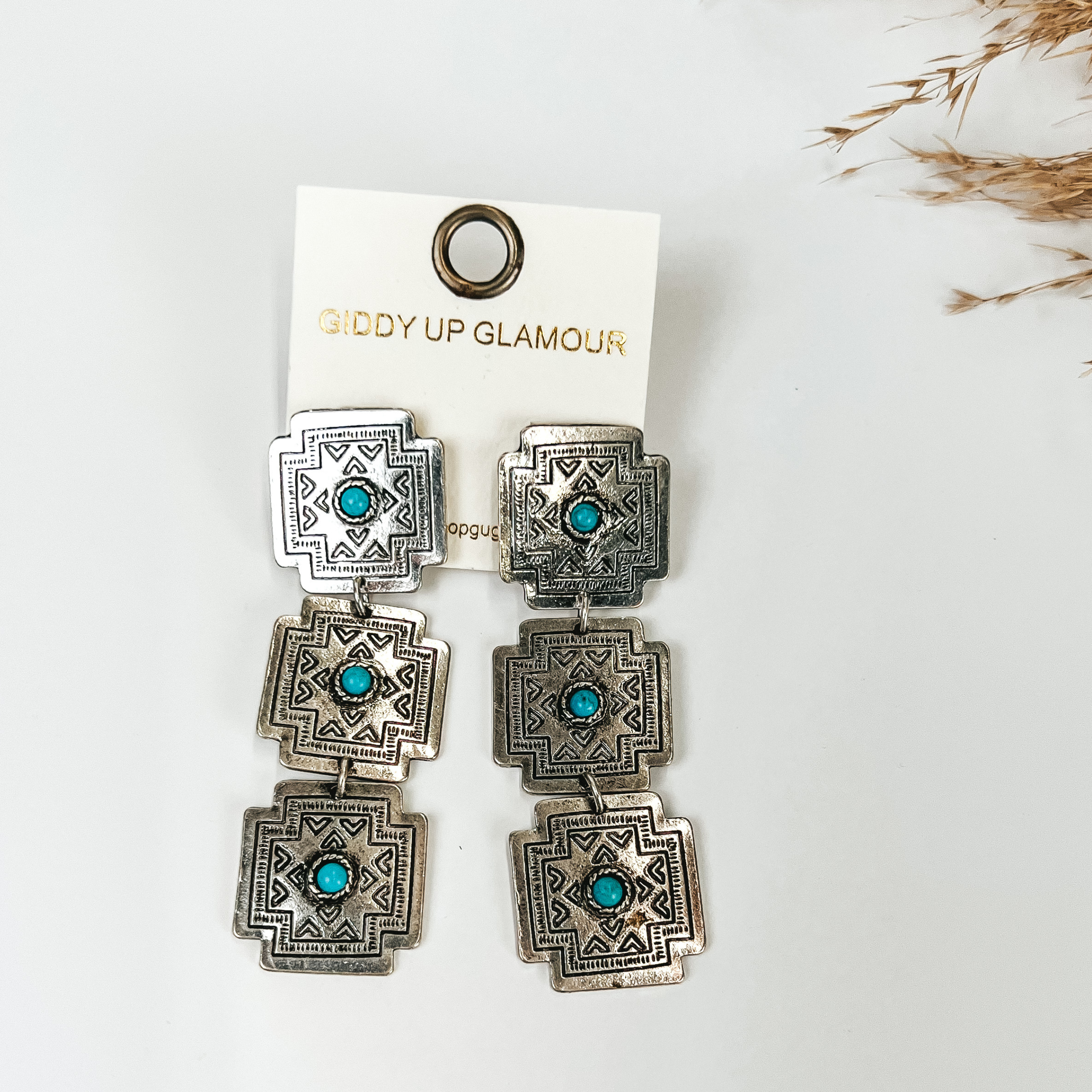 Western design dangle earrings in silver tone with turquoise pendant in the middle, pictured with pampas grass, on a white background. 