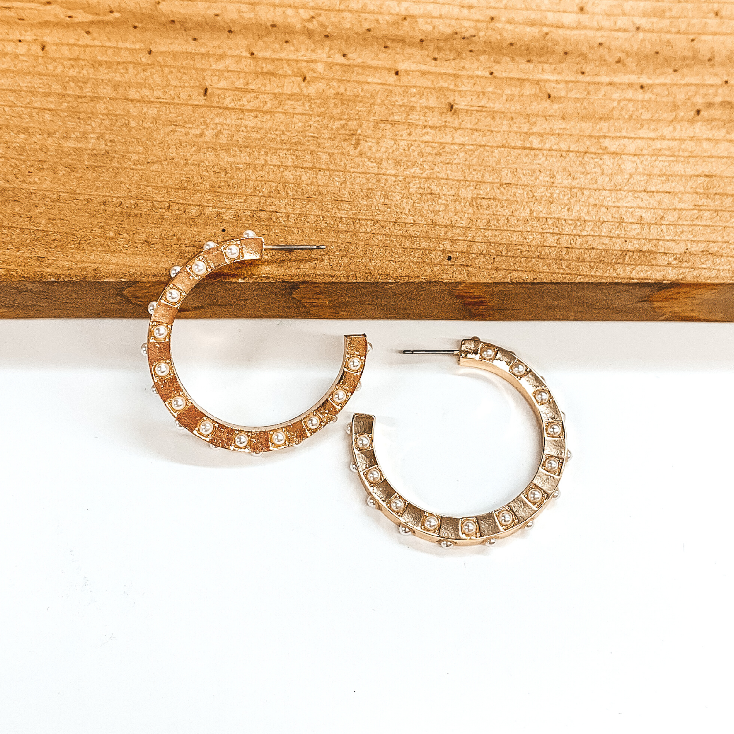 Gold hoop earrings with spaced out white pearls. One earrings is pictured laying on a brown block while the other earrings is pictured laying flat on the white background.