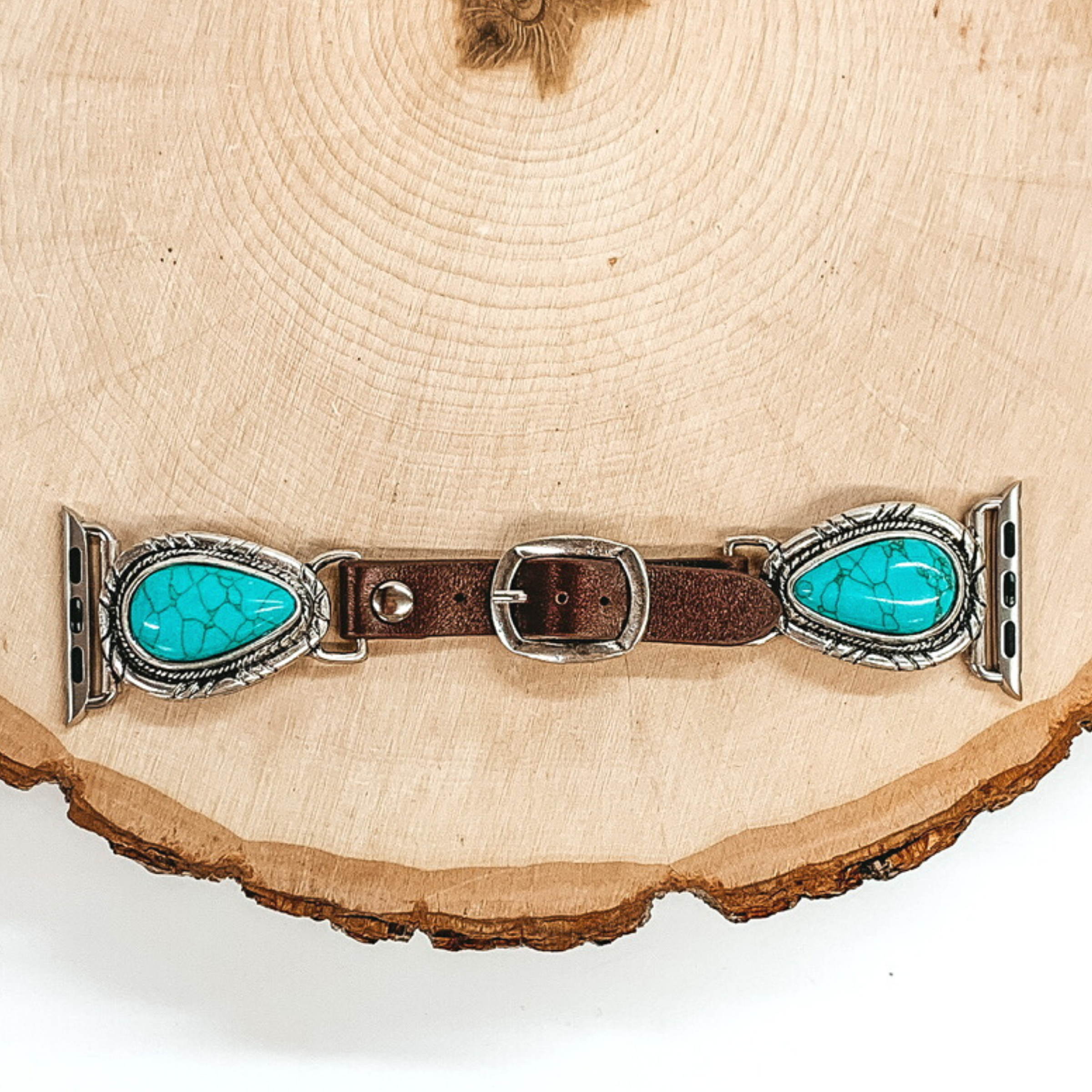 Dark Brown watch band with silver, teardrop pendant ends with Apple watch band acessories. The teardrop pendant has a teardrop shaped turquoise stone. This watch band is pictured on a piece of wood on a white background.