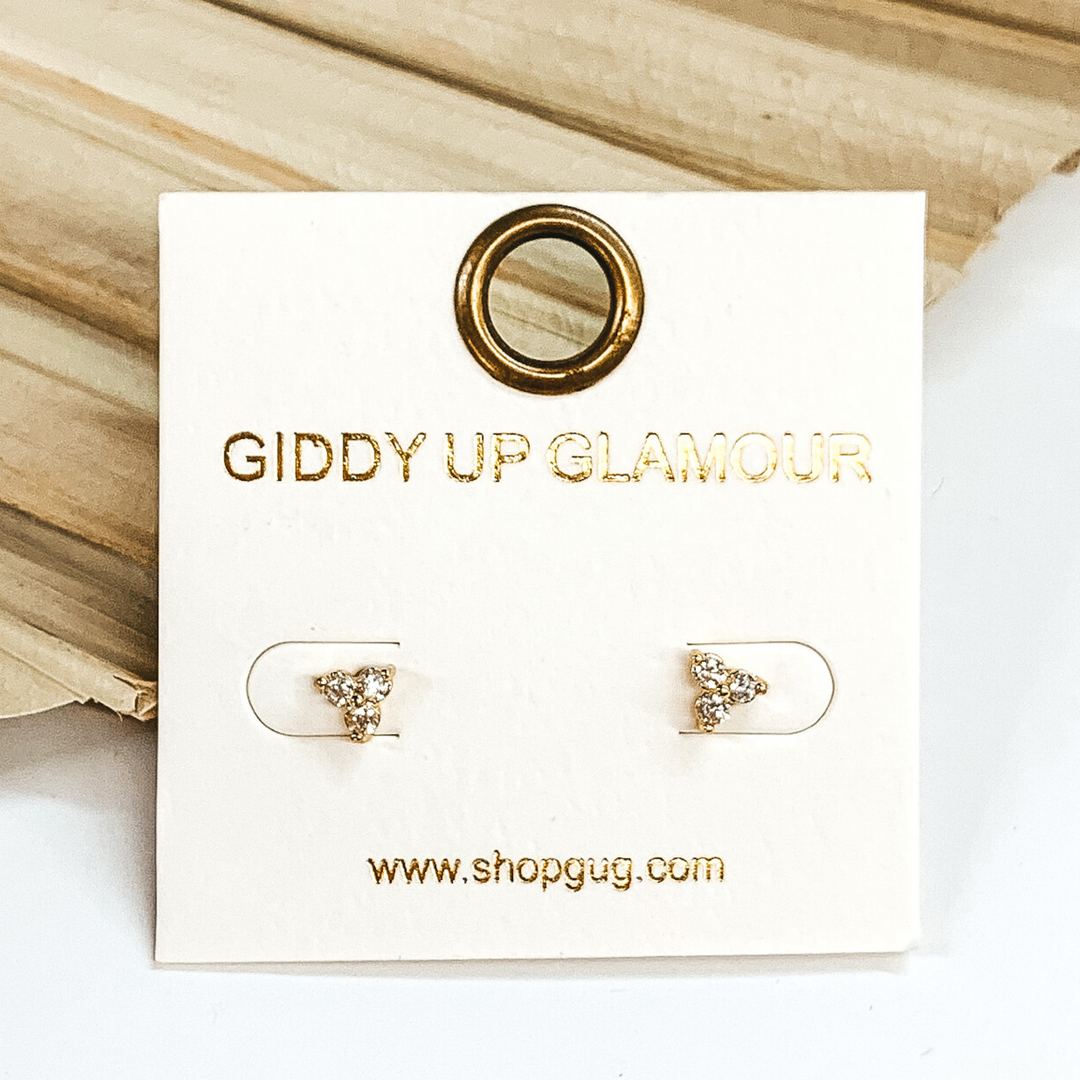 Gold triple crystal stud earrings in a triangle shape in the color gold. These earrings are pictured laying on a green palm leaf on a white background.