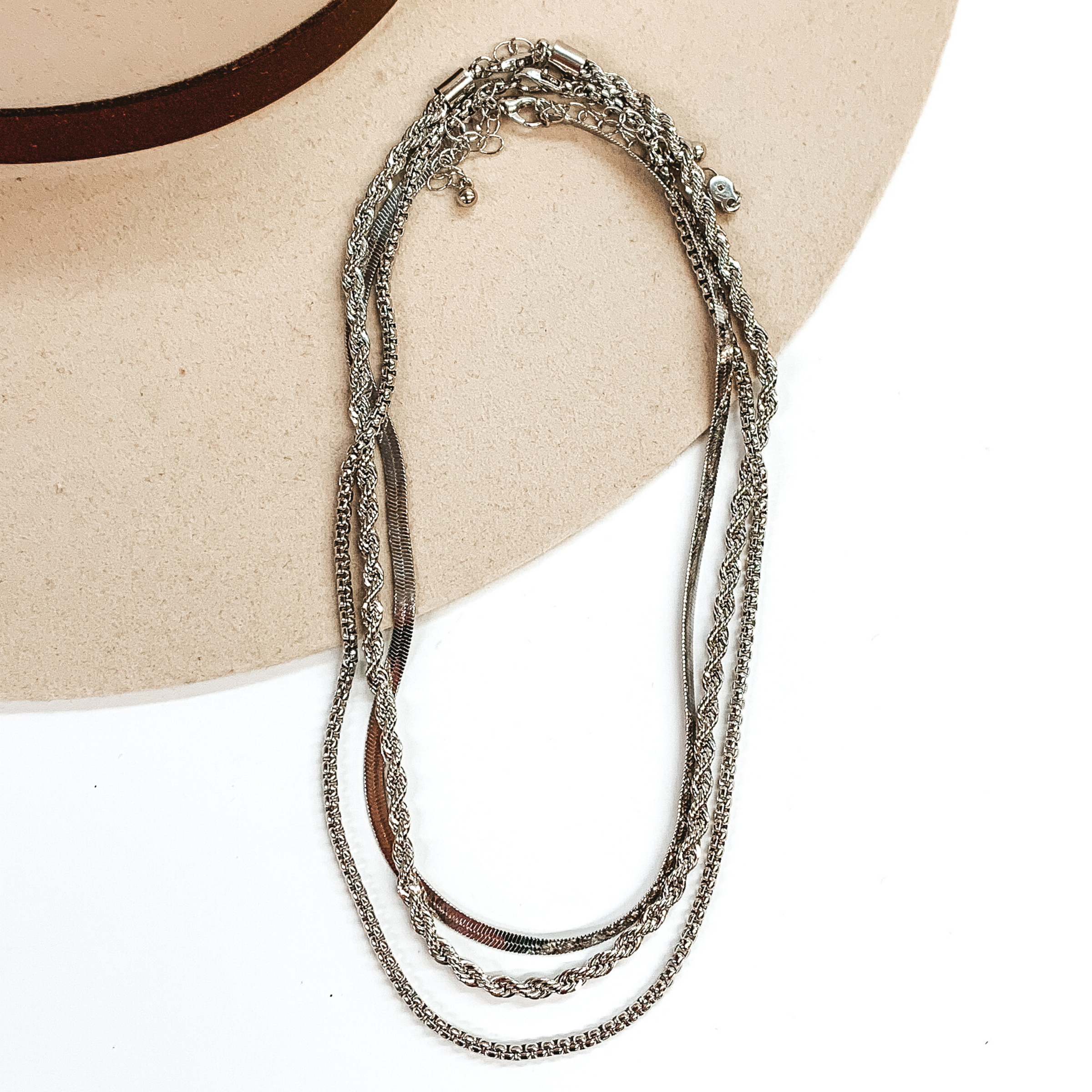 This set of silver necklace include a snake chain, a rope chain, and a box chain necklace. Each necklace has a different length. These necklaces are pictured partially laying on top of a beige hat brim on a white background.