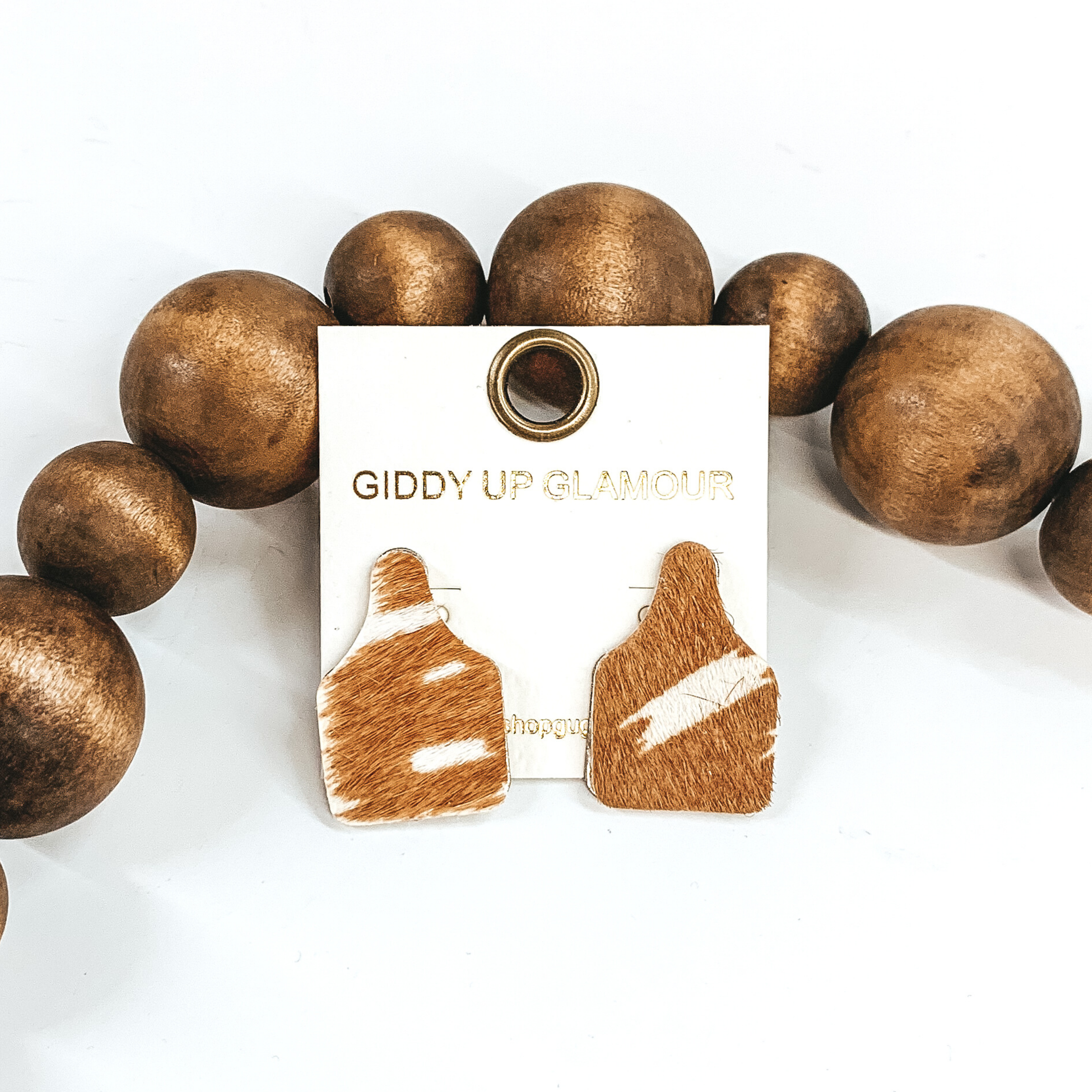 Cattle tag stud earrings with a hair on hide material. It has a cow print in colors tan and white. These earrings are pictured in front of dark brown beads on a white background. 