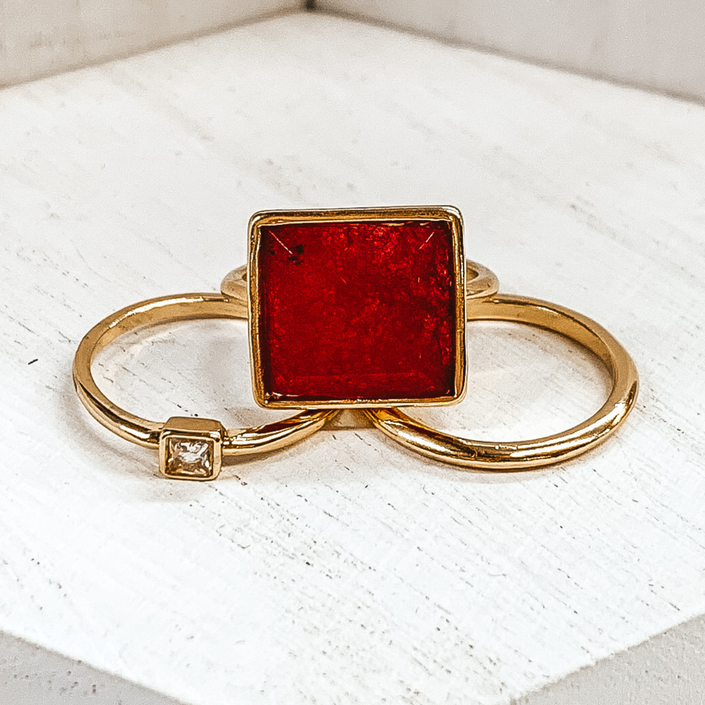 Set of three gold rings. One ring is a plain gold band. Another has a small square pendant with a center crystal. The third ring has a big, maroon pendant. These rings are pitured on a white background.  
