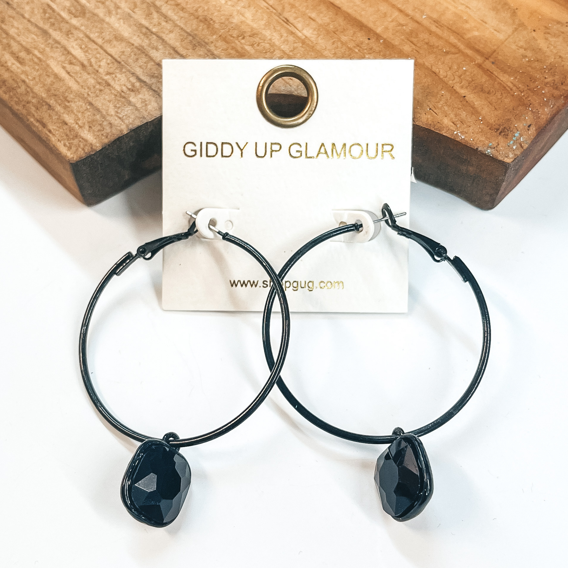 Circle hoop earrings in black with a cushion cut black crystal drop. Taken on a white background and leaned up against  a brown block.
