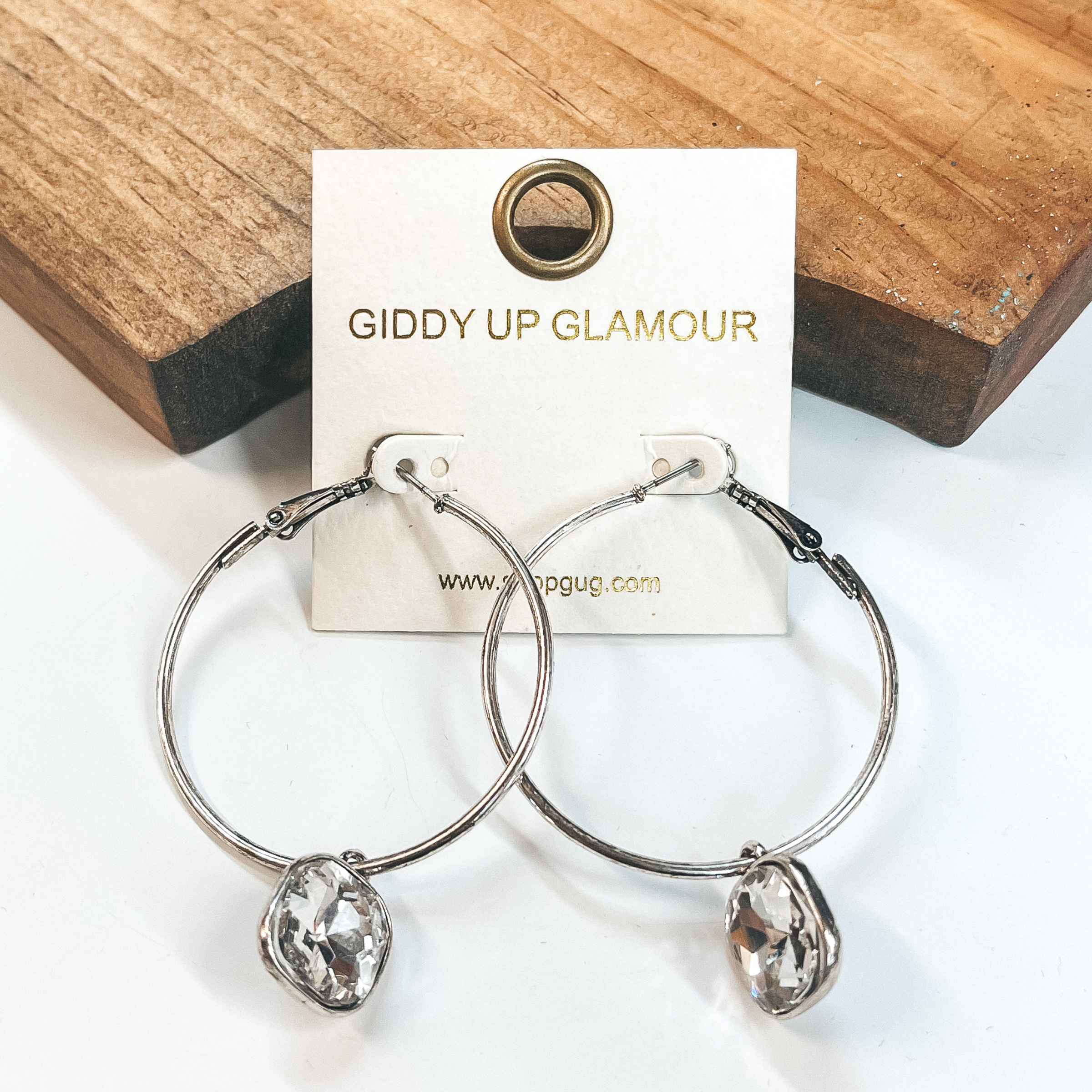Circle hoop earrings in silver with a cushion cut clear crystal drop. Taken on a white background and leaned up against  a brown block.