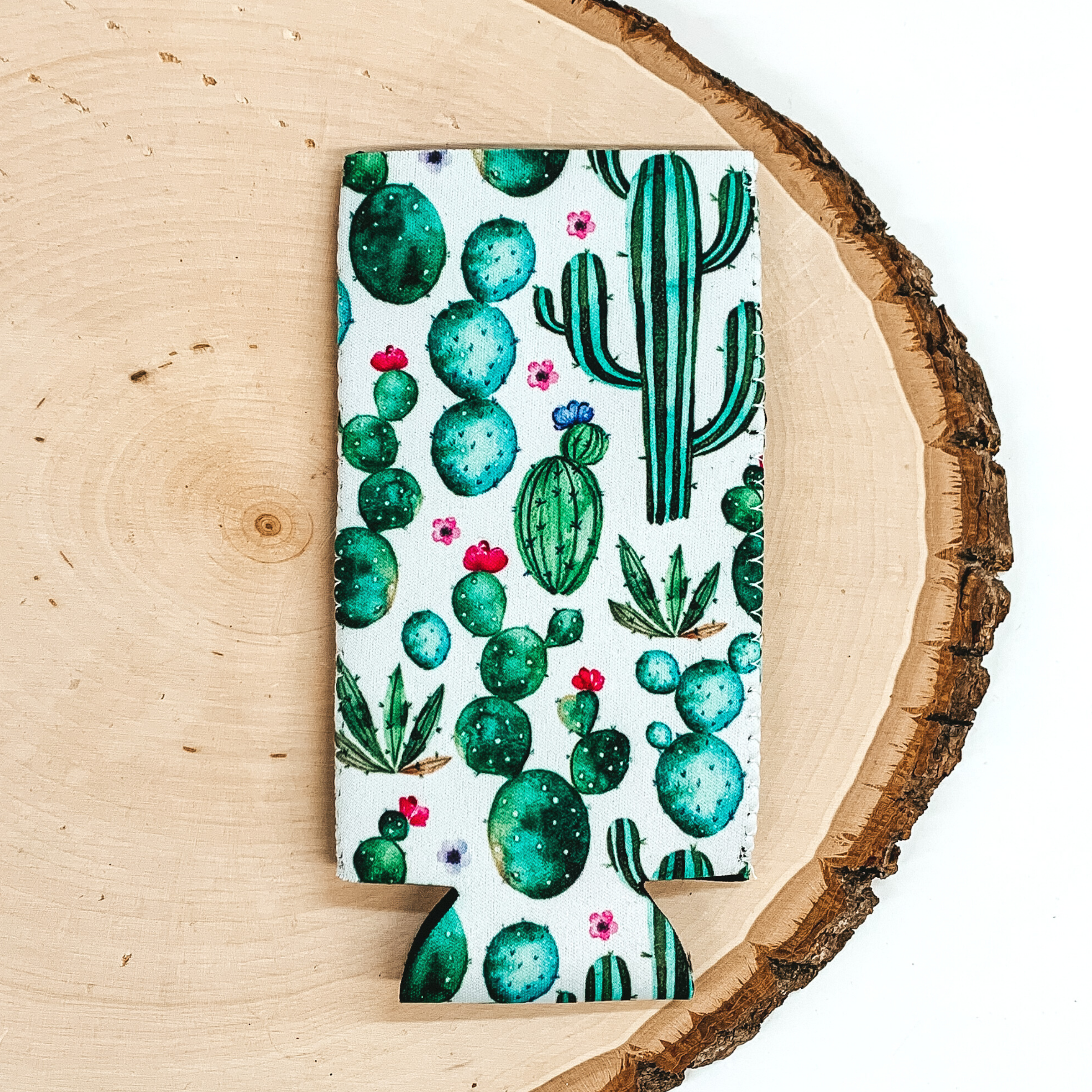 White slim can koozie with different shaped cacti in different shades of green and a few pink flowers. This koozie is pictured laying on a piece of wood on a white background.