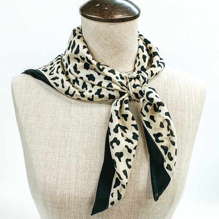 Tan scarf with black leopard print and black outline tied around the neck of a mannequin. Pictured on white background.