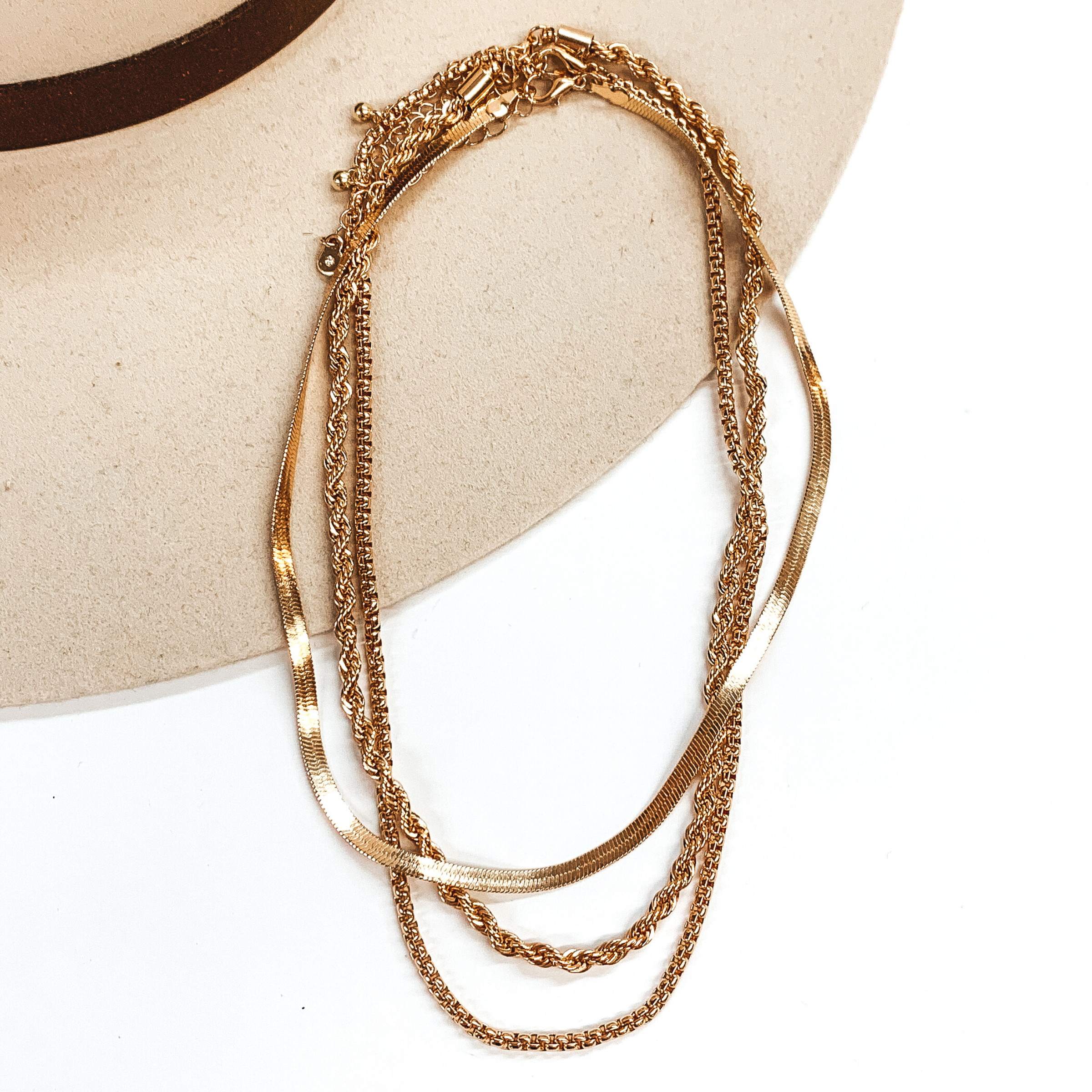 This set of gold necklace include a snake chain, a rope chain, and a box chain necklace. Each necklace has a different length. These necklaces are pictured partially laying on top of a beige hat brim on a white background.